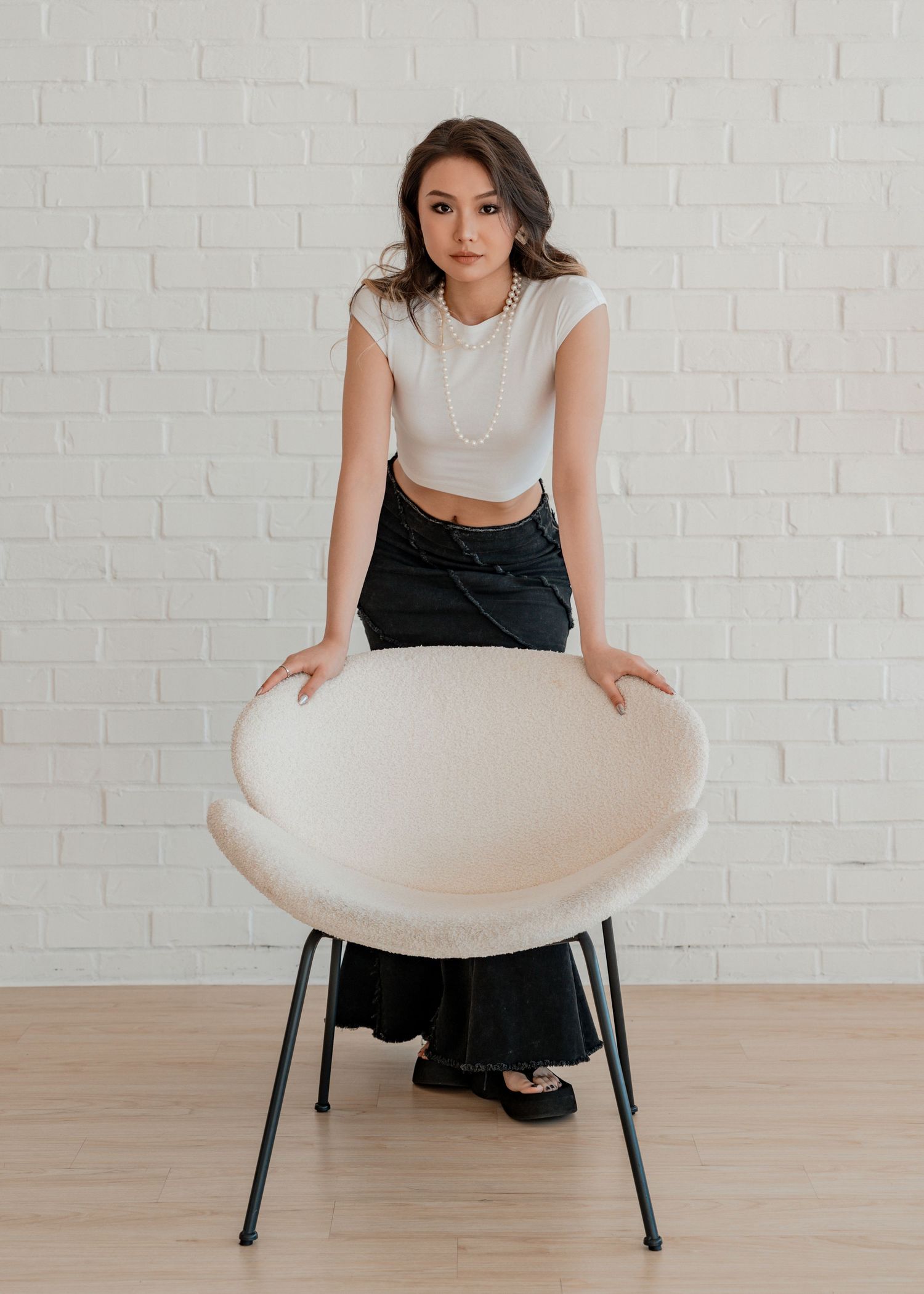 Elegant young woman in white crop top leaning on round cream accent chair in modern studio.