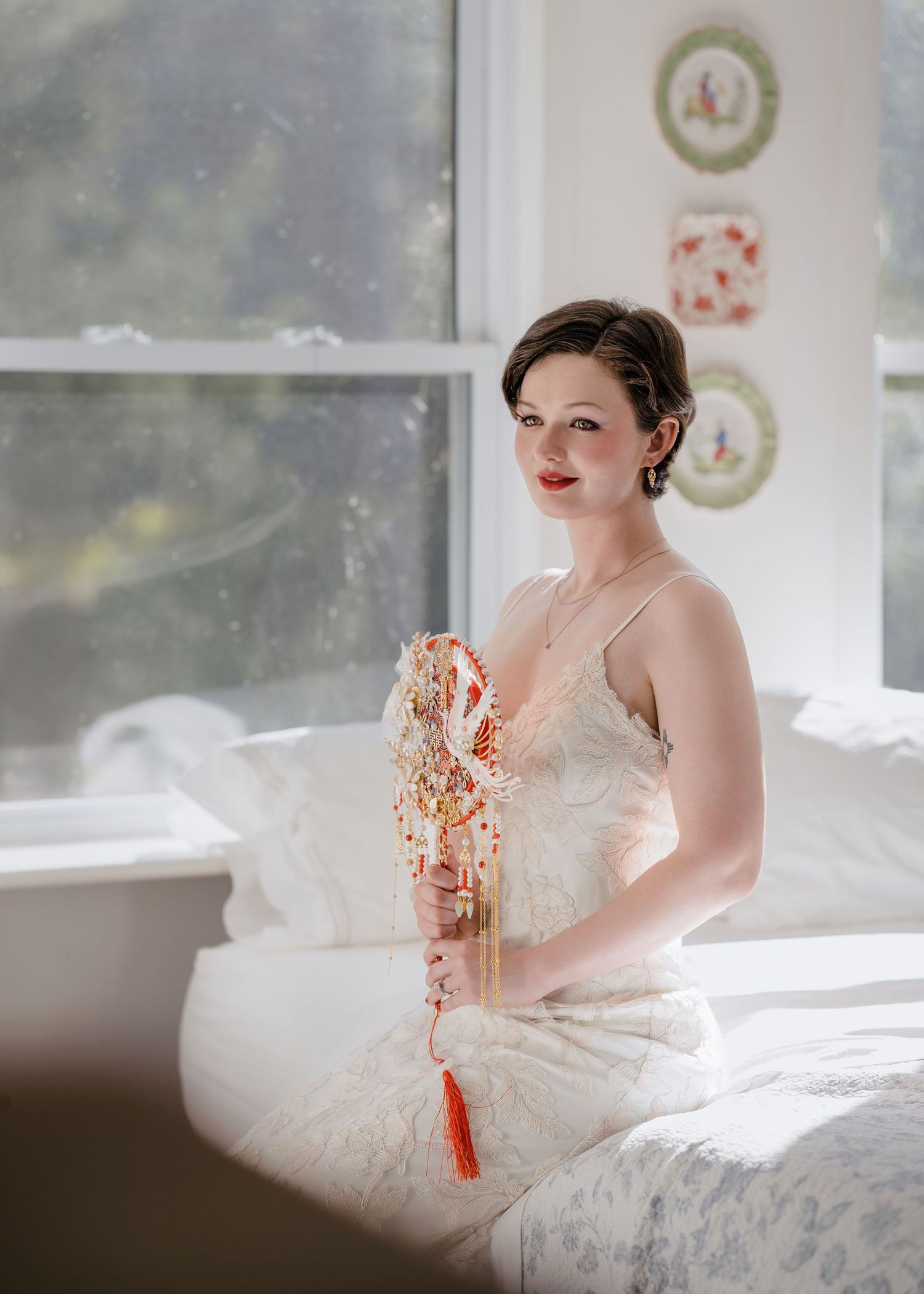 Beautiful bride in lace gown sitting on bed holding colorful beaded bouquet by sunny window.