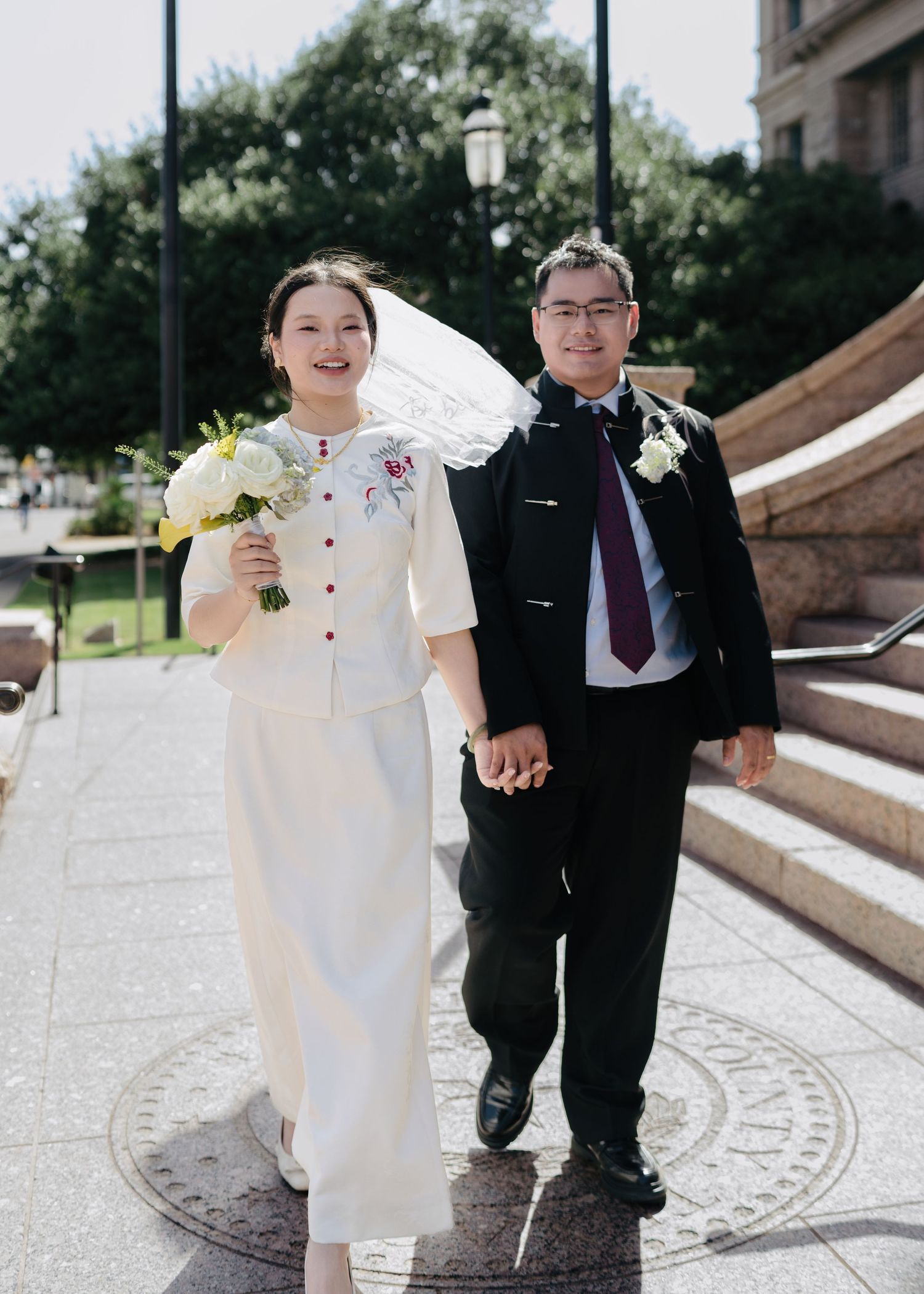 Happy couple in wedding attire holding hands outdoors, bride in white dress with veil holding bouquet, groom in black suit.
