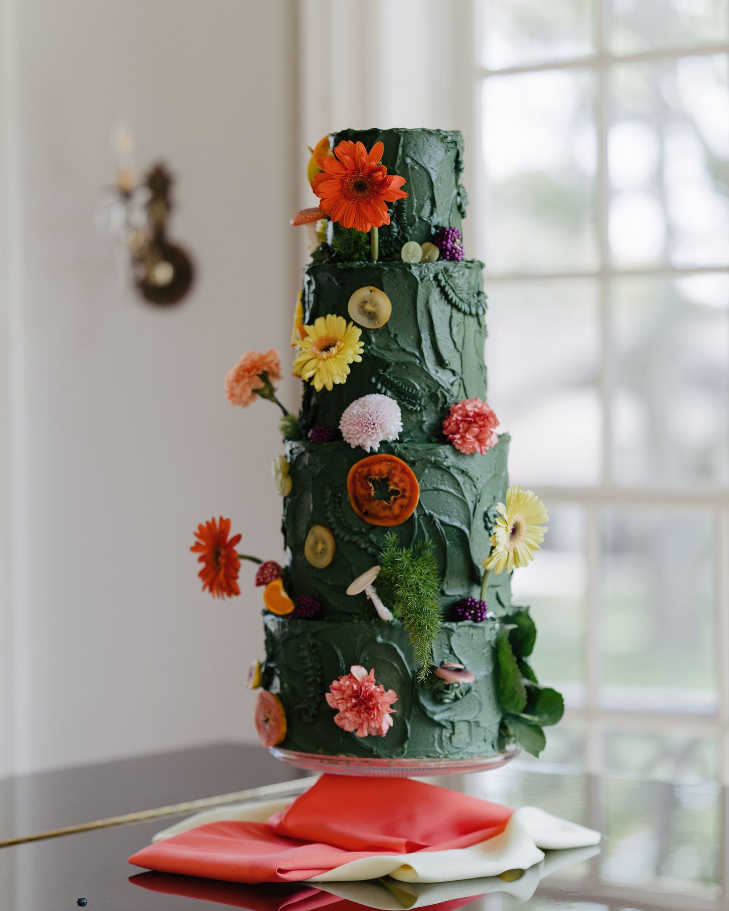 Stunning four-tier dark green wedding cake adorned with colorful fresh flowers and textured buttercream.