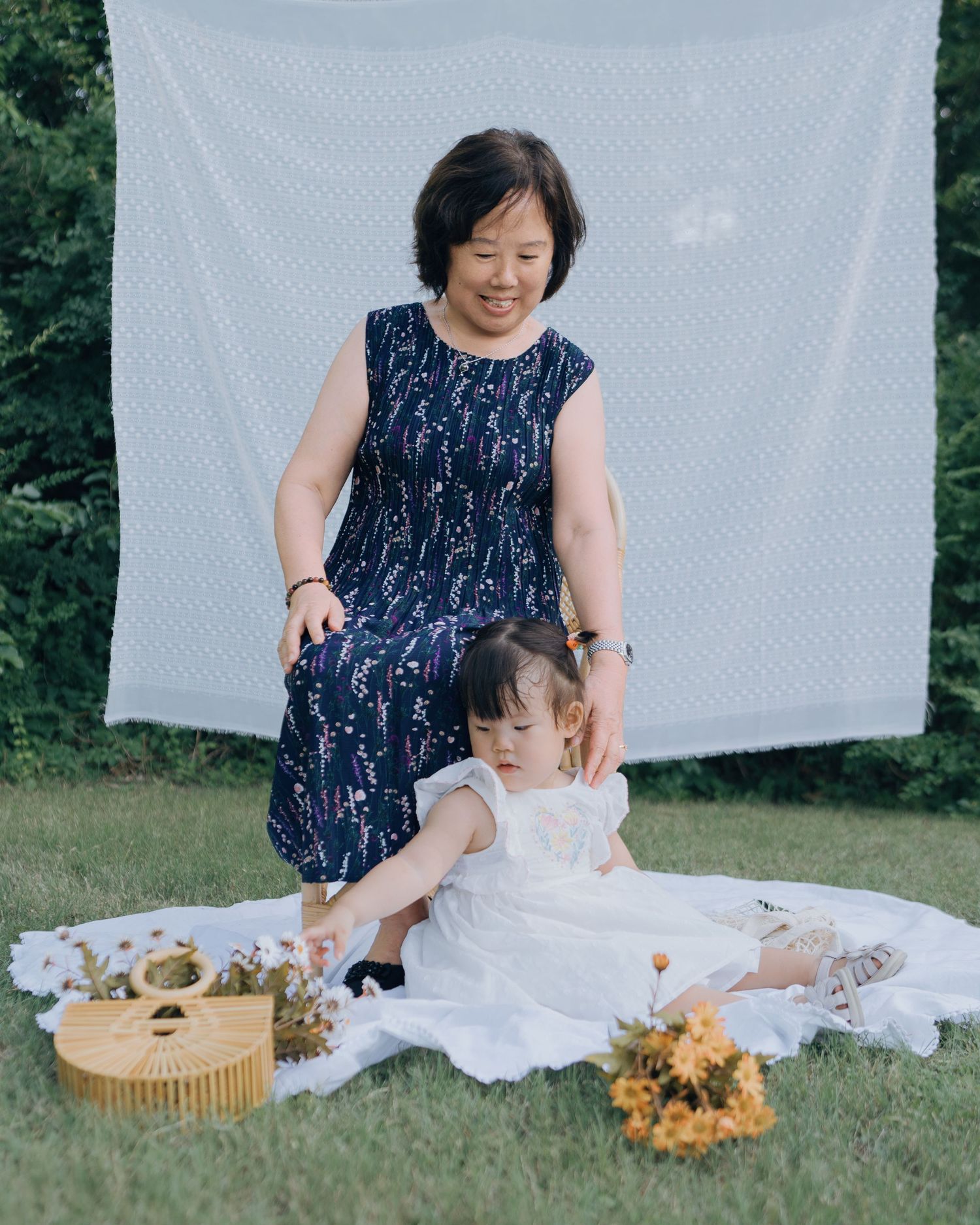 Grandmother and toddler girl playing on white blanket in garden with yellow flowers and wicker basket.
