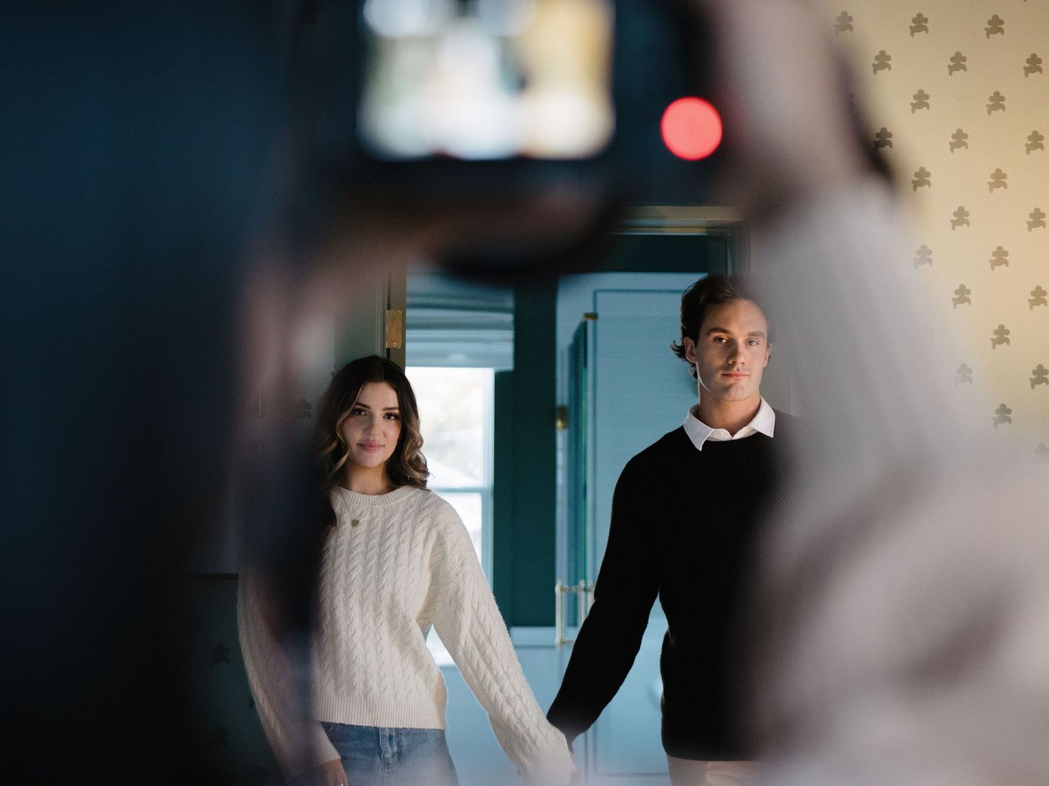 Couple holding hands being photographed by photographer in a bright room with patterned wallpaper.