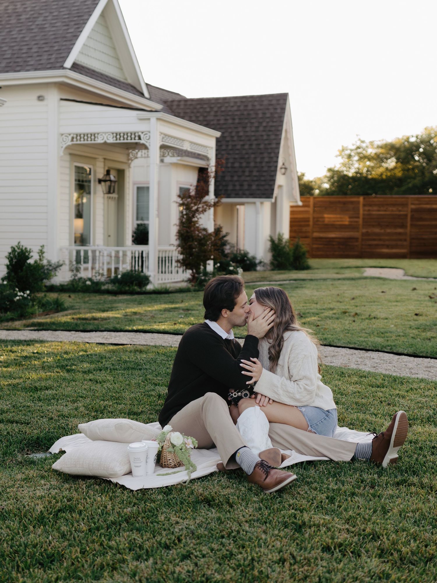 Couple kissing while seated on a blanket on the lawn in front of a white cottage.