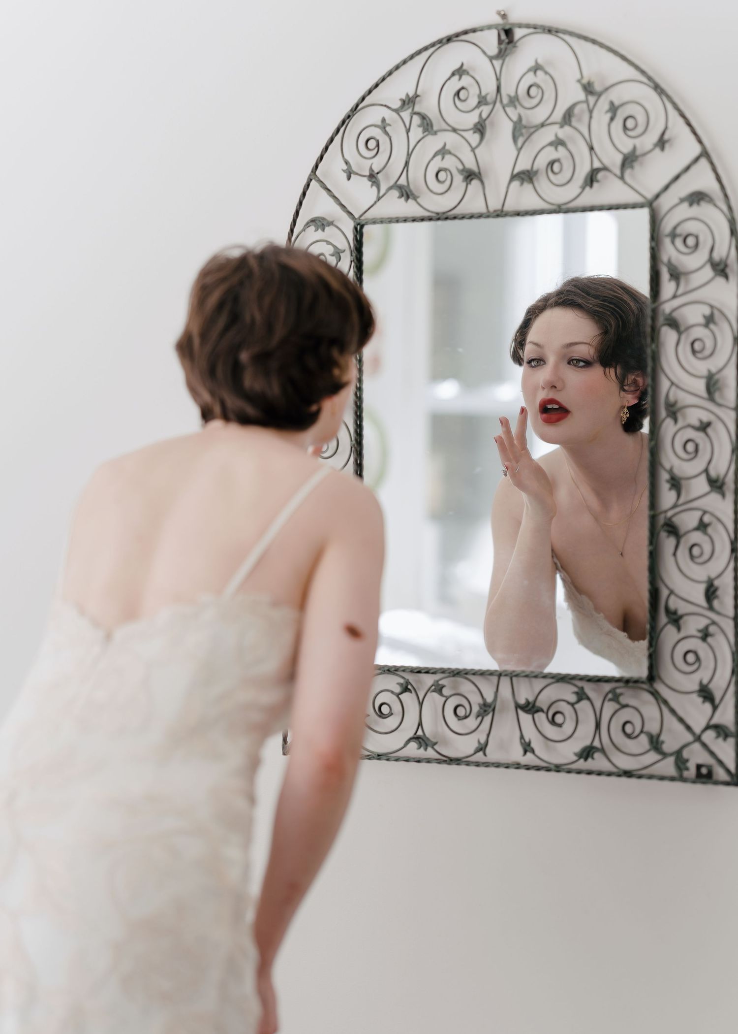 Bride in lace wedding dress checking red lipstick in ornate wrought iron mirror.