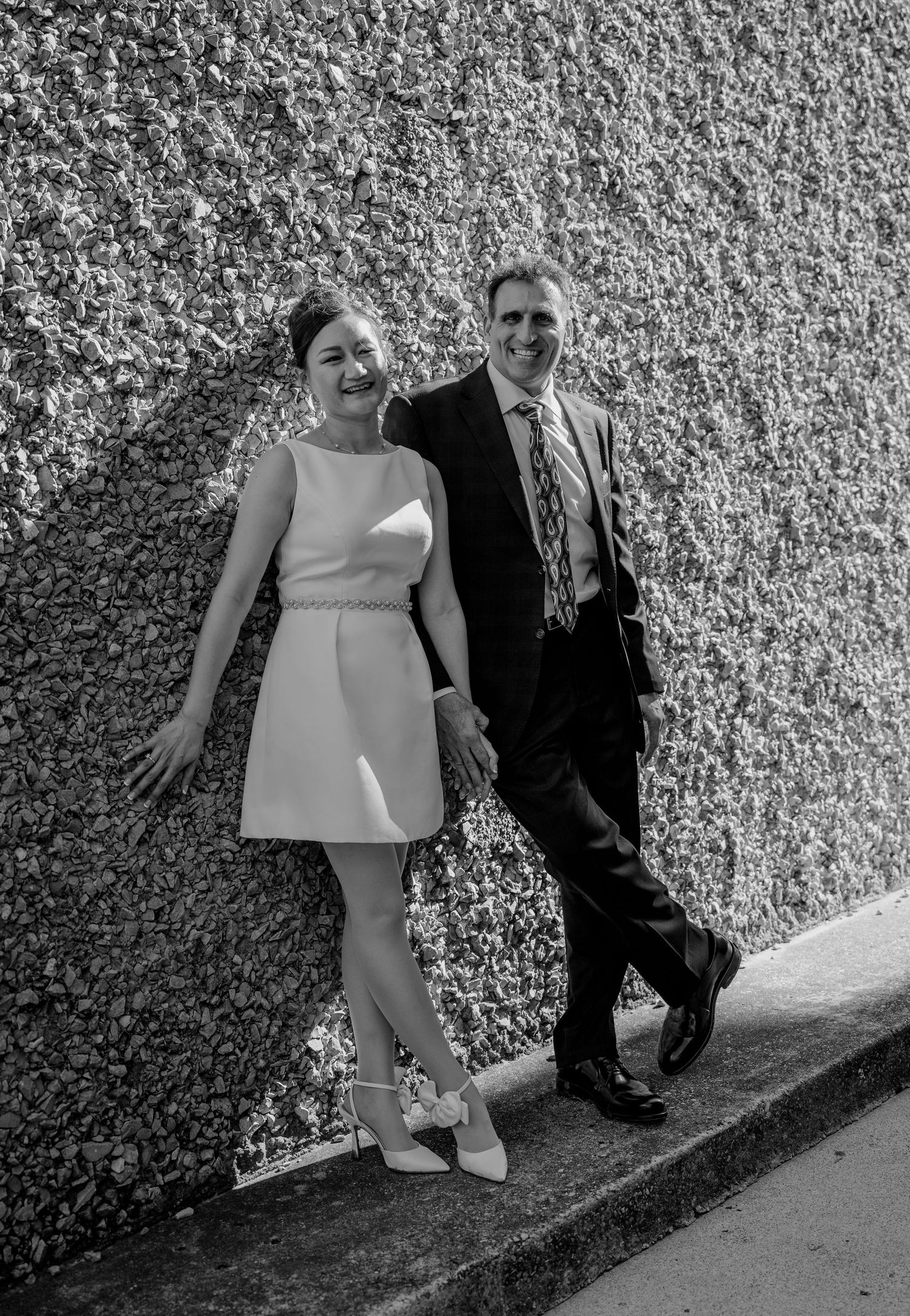 Black and white photo of smiling couple leaning against ivy-covered wall outdoors.