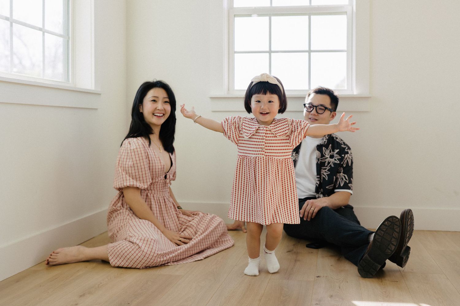 A happy Asian family with a toddler girl in pink stripes spreading her arms wide in a bright room.