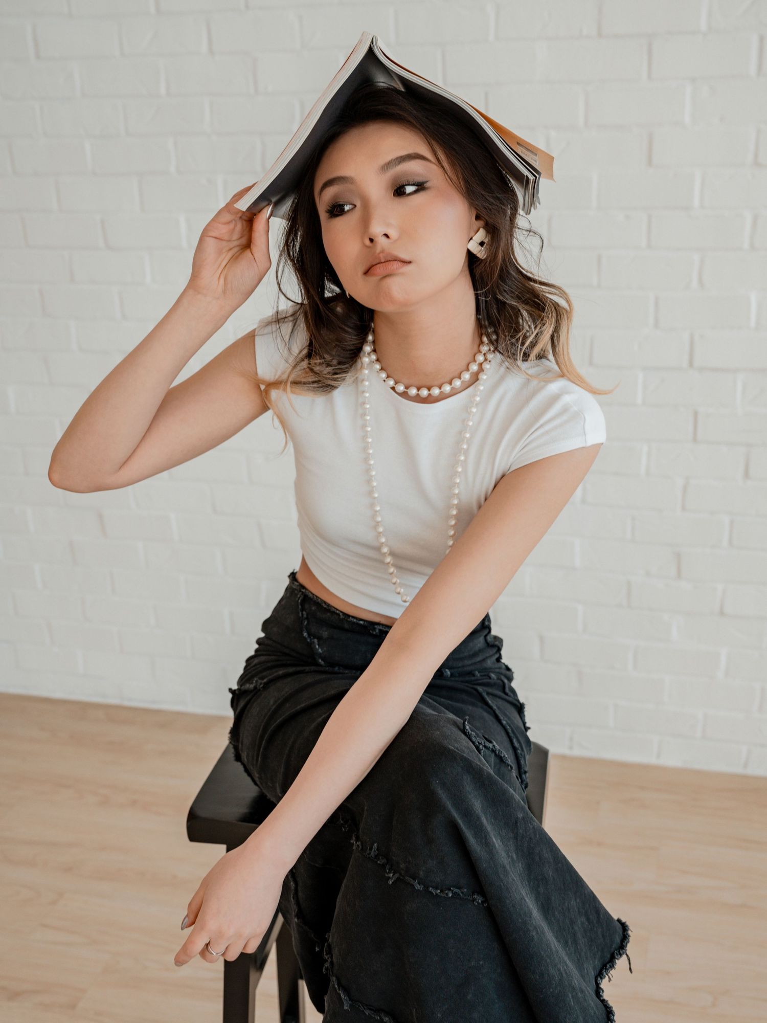 Young woman in white crop top and pearl necklace balances an open book on her head while seated.
