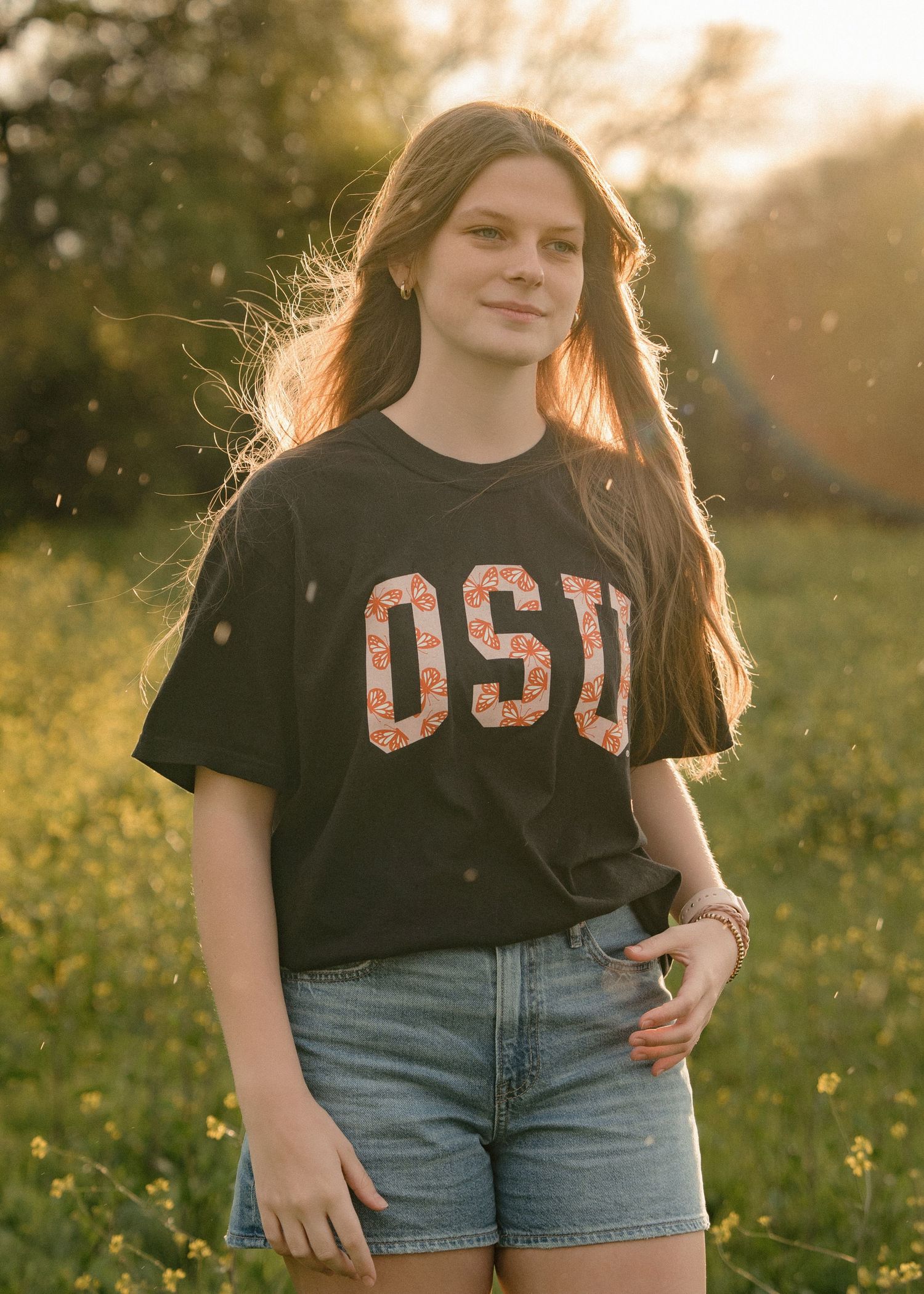 Teen girl in black OSU graphic tee and denim shorts smiles in a golden-hour wildflower meadow.