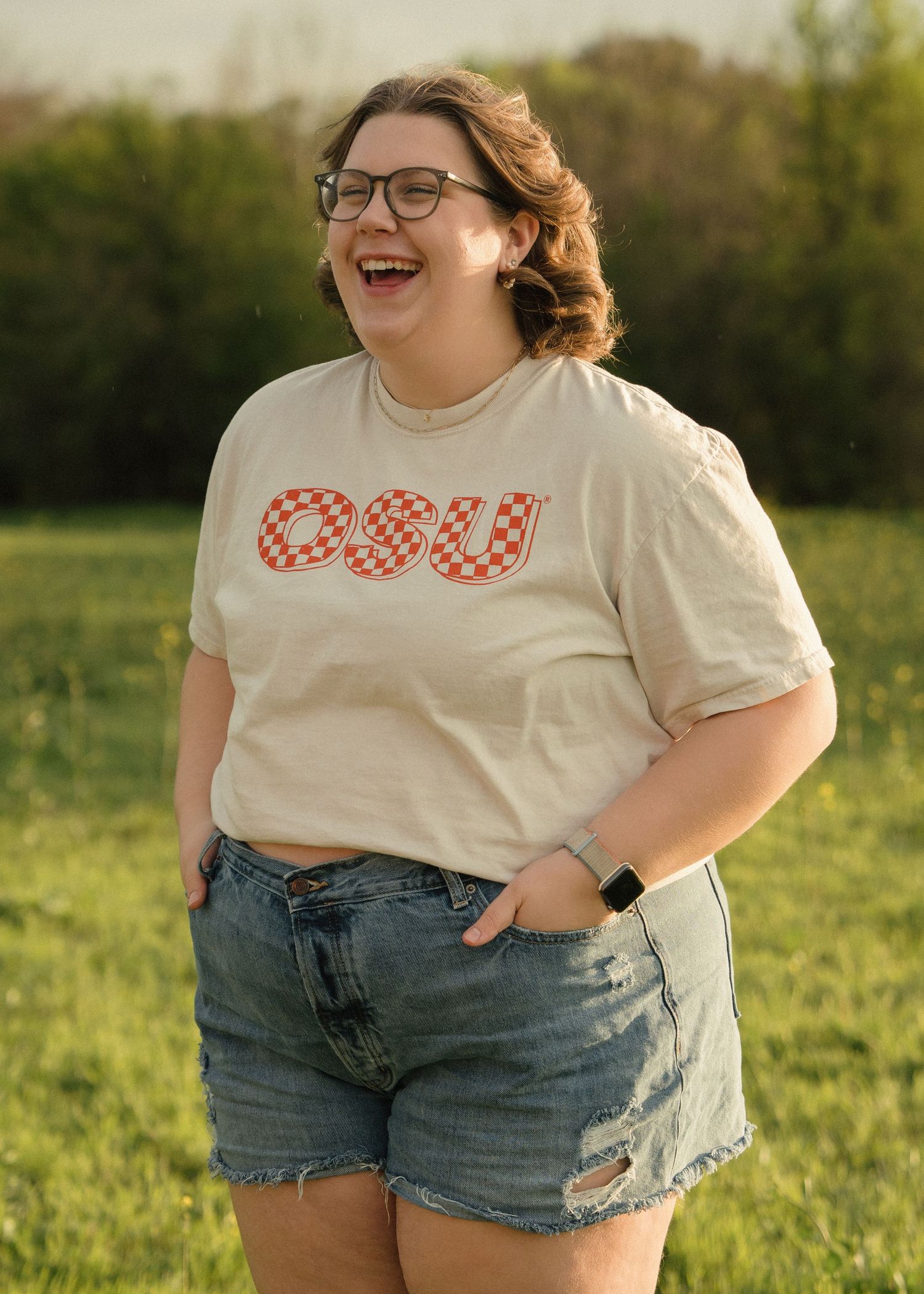 Smiling woman in beige OSU checkered graphic tee and distressed denim shorts standing in green field.