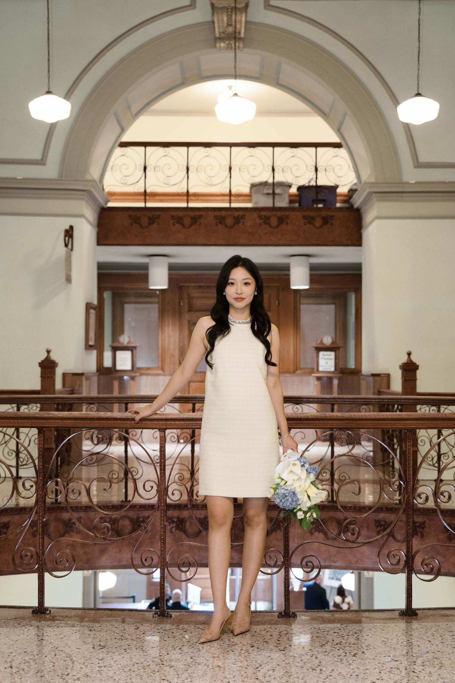 Woman in white halter dress holding hydrangea bouquet standing inside elegant courthouse interior.