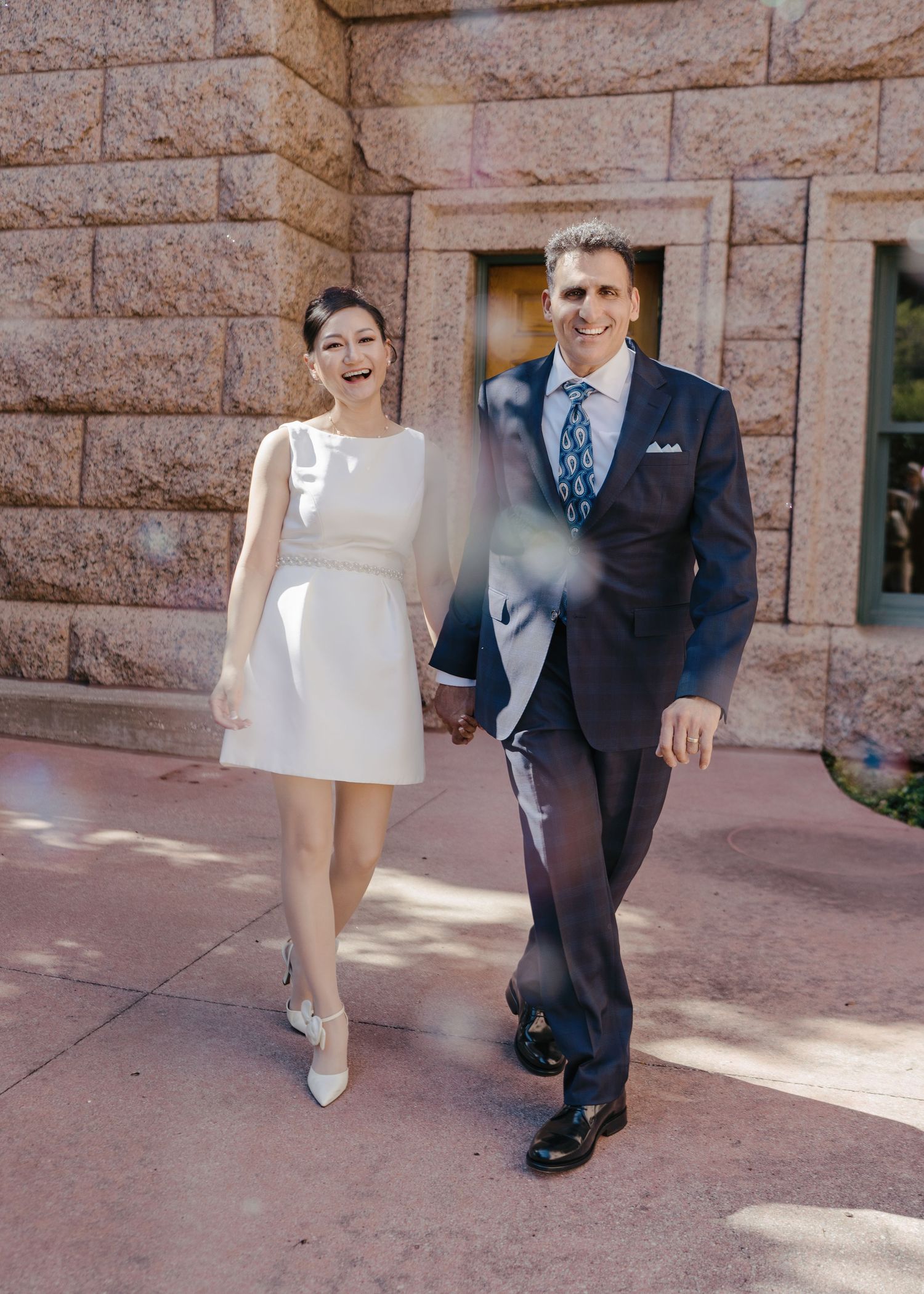 Laughing bride in white mini dress and groom in navy suit exit courthouse after wedding.