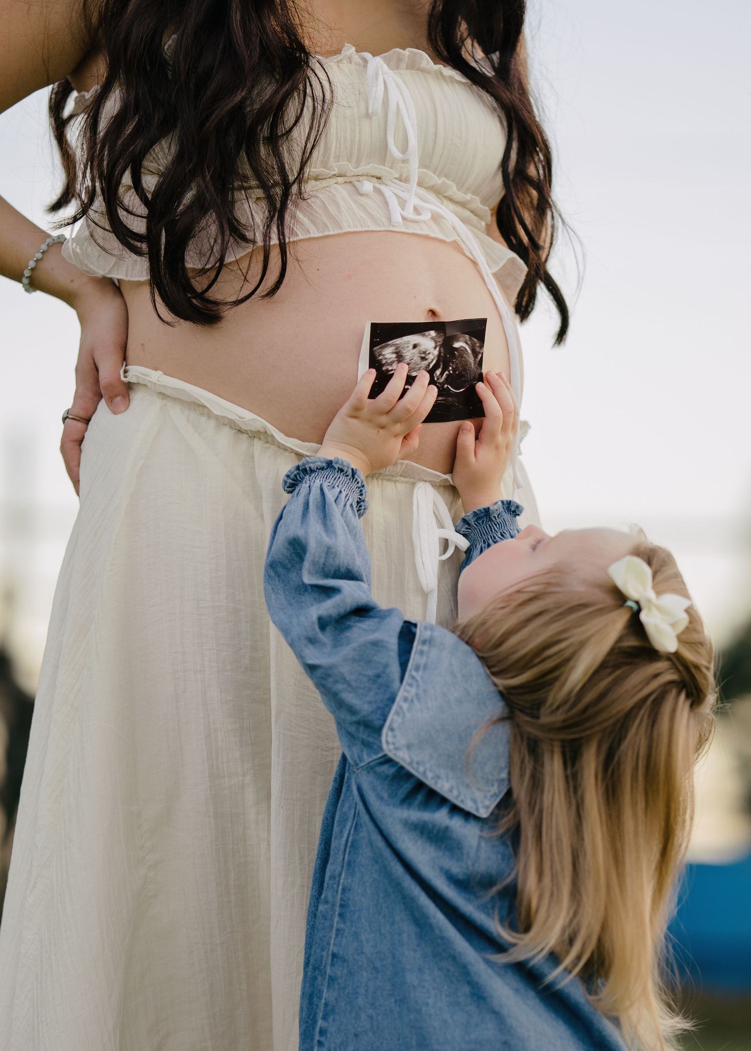 Young blonde girl holding ultrasound photo against pregnant mothers belly in white maternity dress.