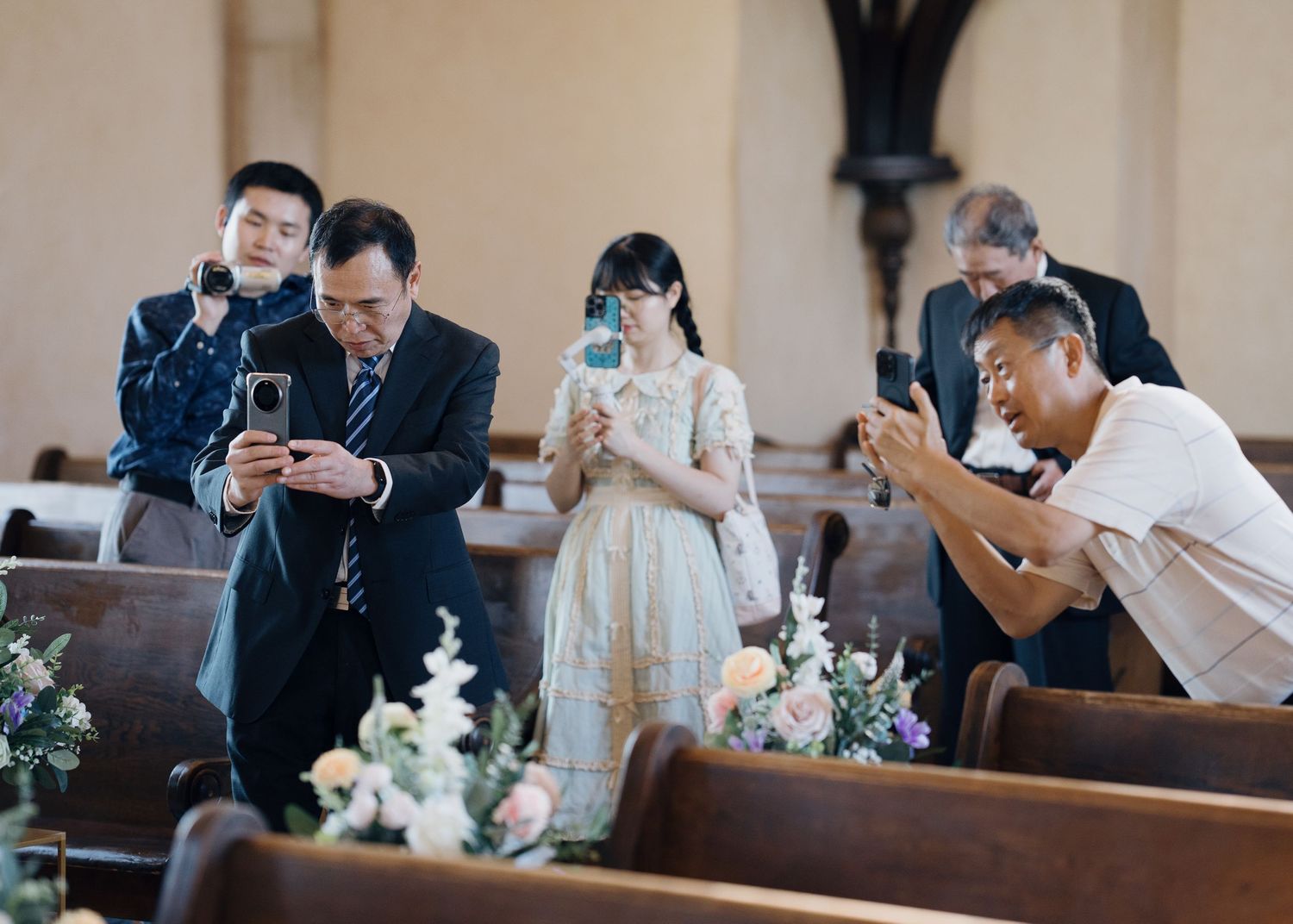 Wedding guests photographing a bride in a church aisle surrounded by floral arrangements on golden stands.
