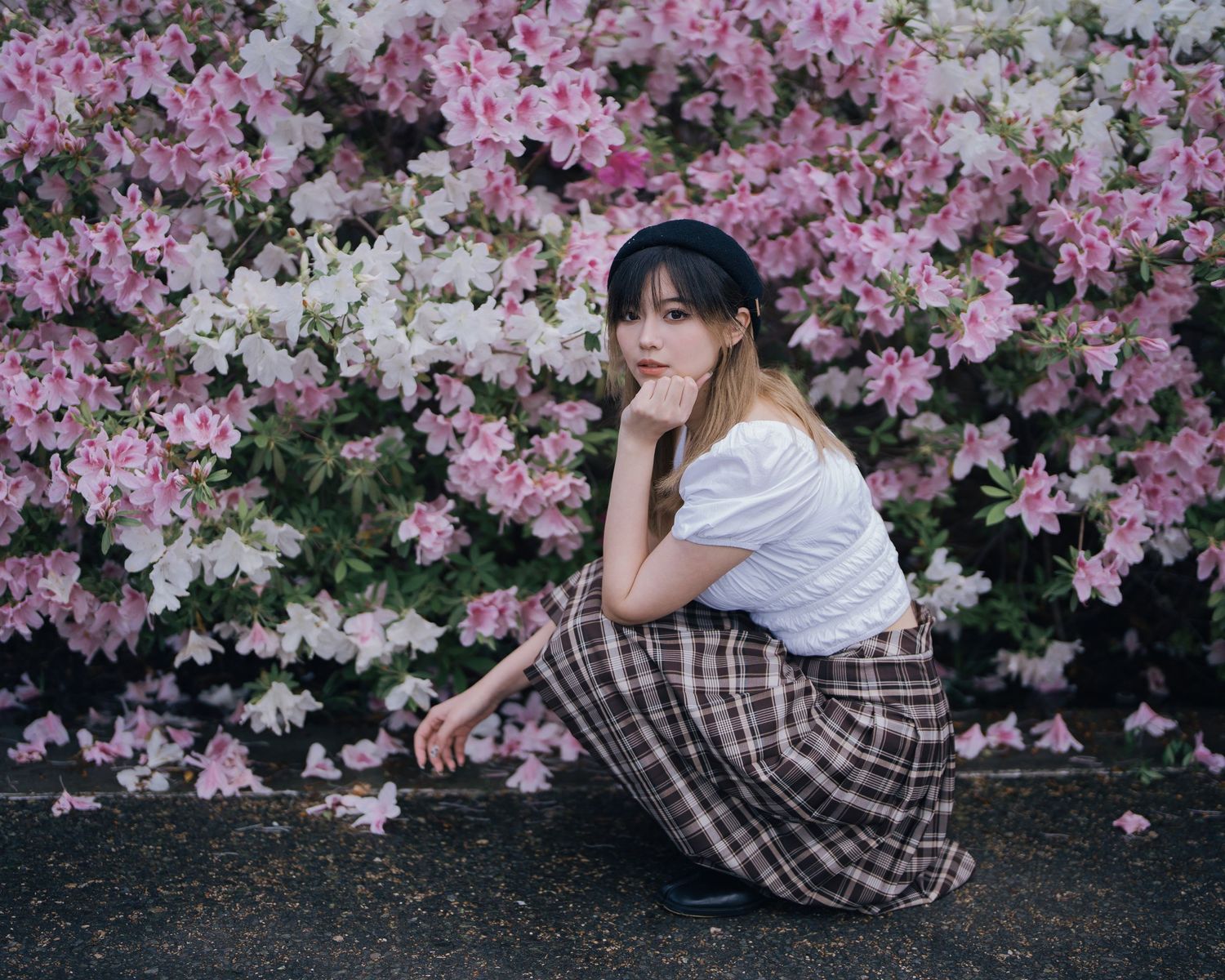 Young Asian woman in plaid skirt and white top posing beside pink and white azalea bushes.