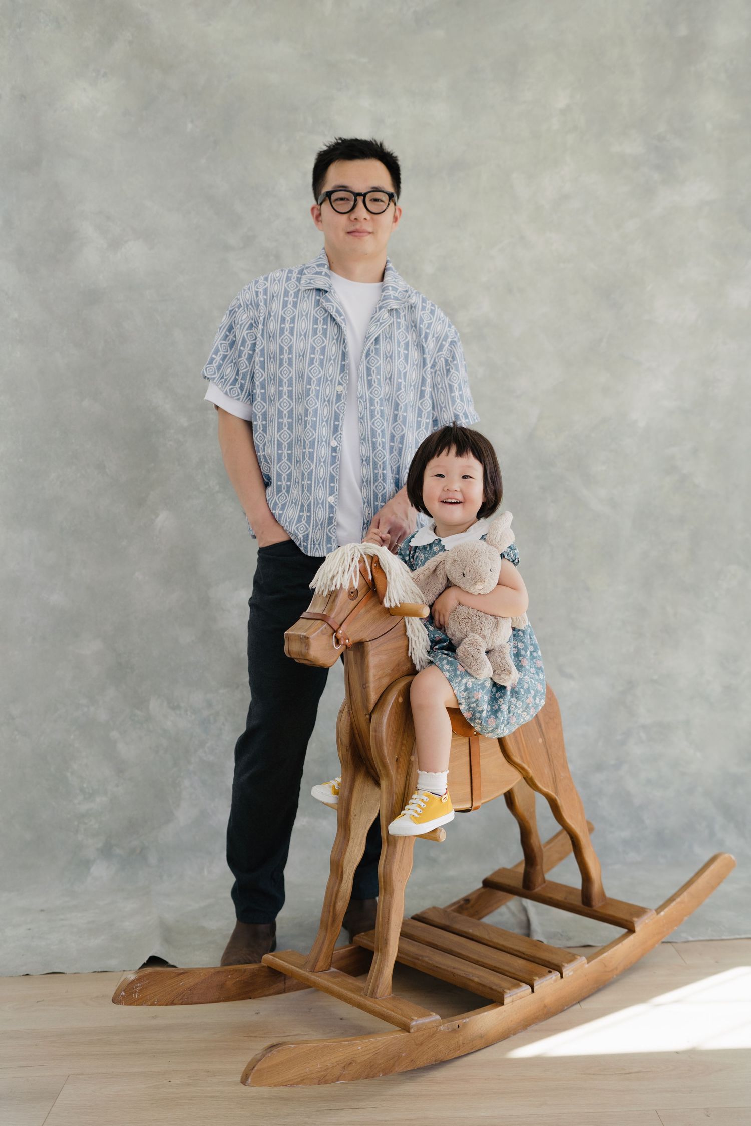 Father standing beside smiling toddler daughter sitting on wooden rocking horse with stuffed animal.