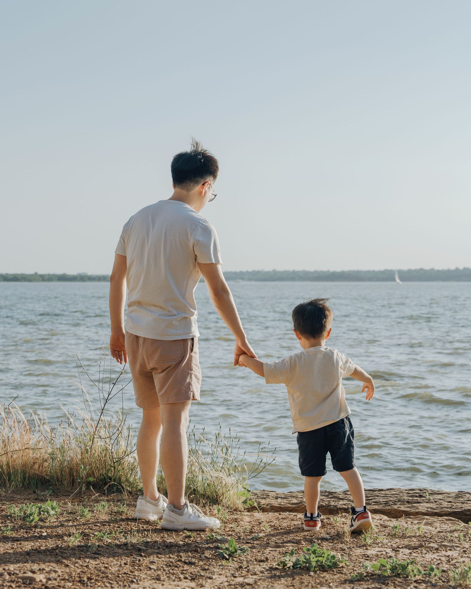 Father and young son hold hands while standing at the edge of a calm lake on a sunny day.