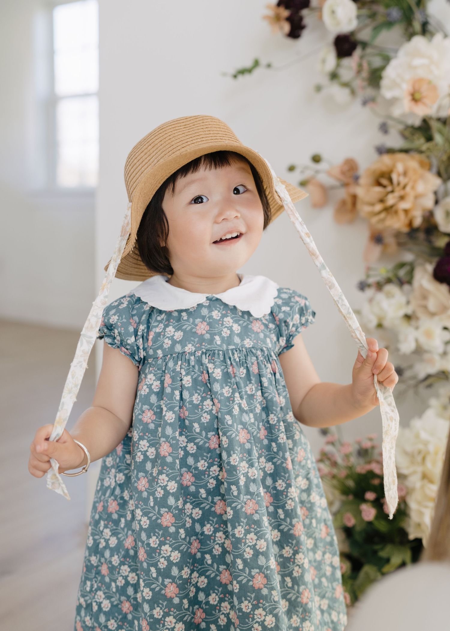 Cute toddler girl in teal floral dress and straw hat smiling near floral arrangement.