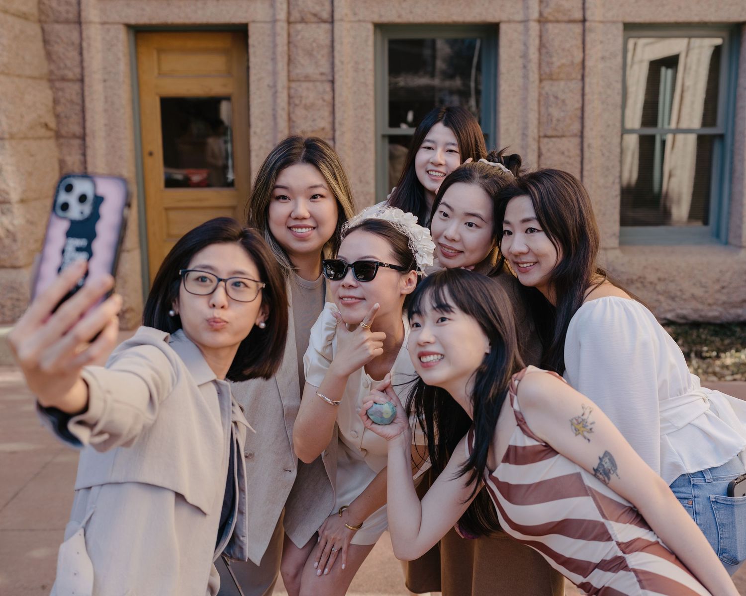 Seven young Asian women pose together for a fun selfie outdoors near a brick building on a sunny day.