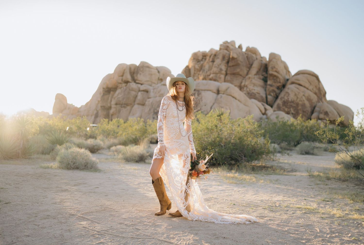 Boho bride in lace dress and cowboy boots walking through Joshua Tree desert landscape at golden hour.