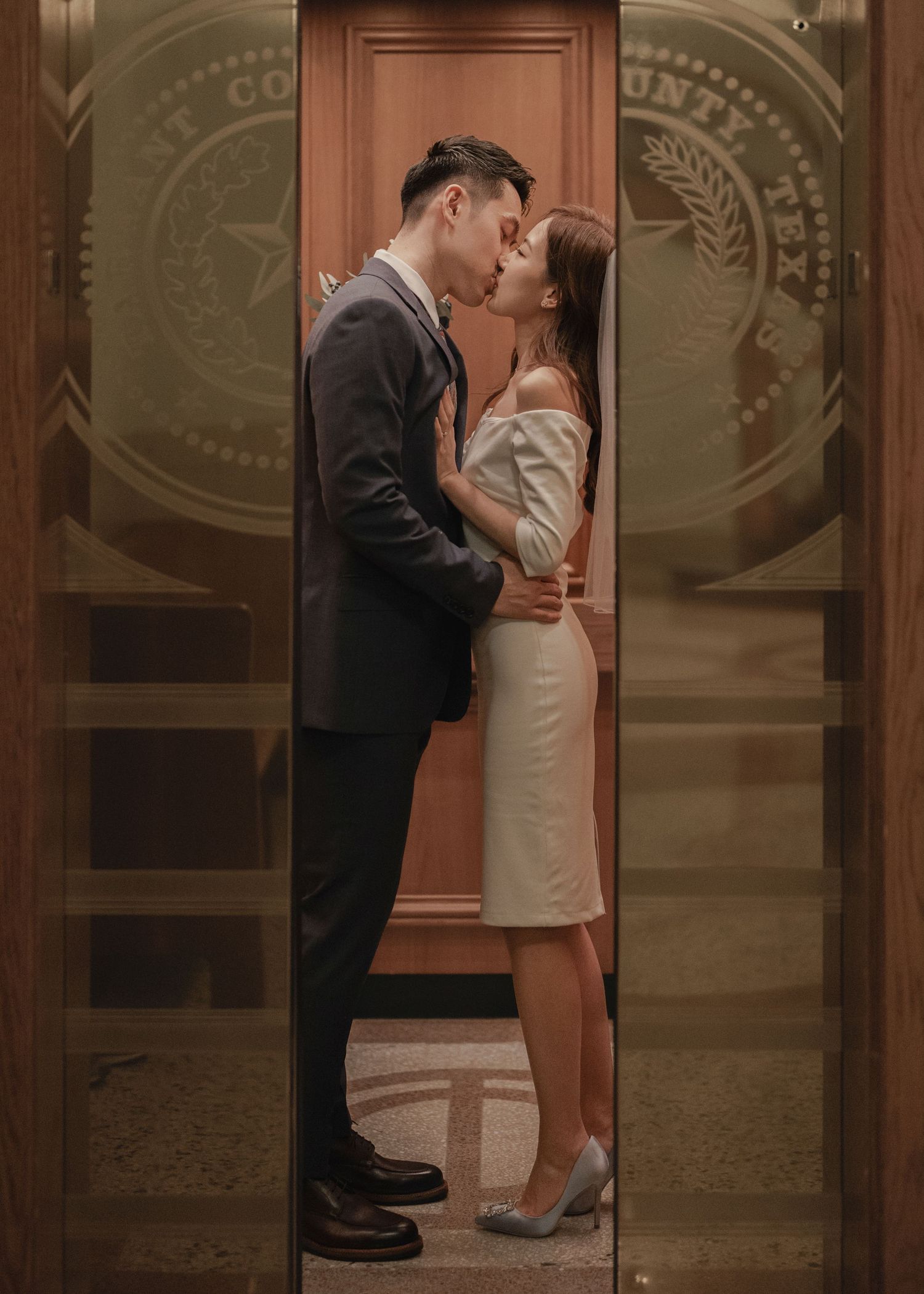 Asian couple sharing romantic kiss in elevator doorway with county seal engraved on door.