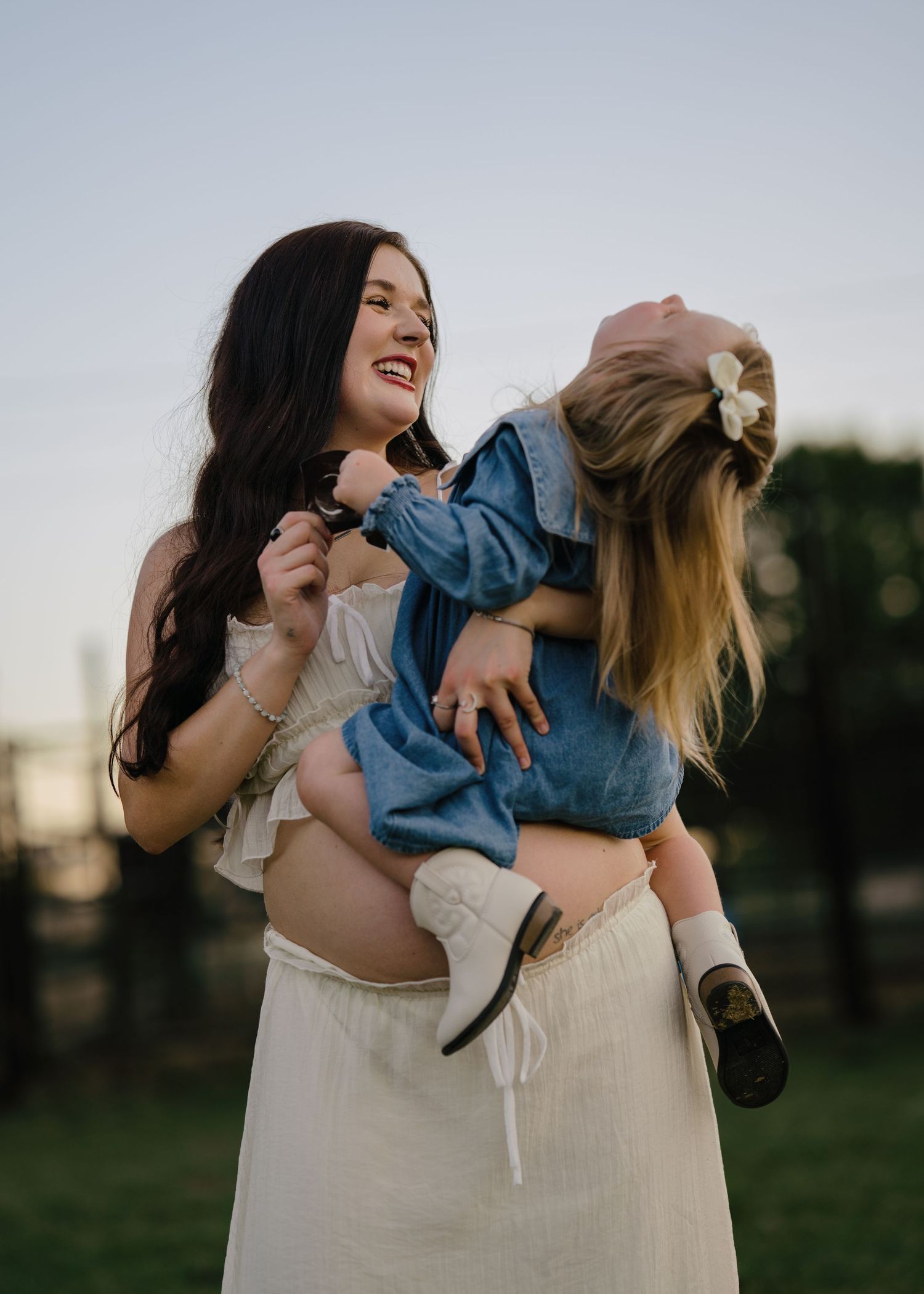Joyful pregnant mother lifting and playing with laughing blonde toddler daughter at sunset outdoors.