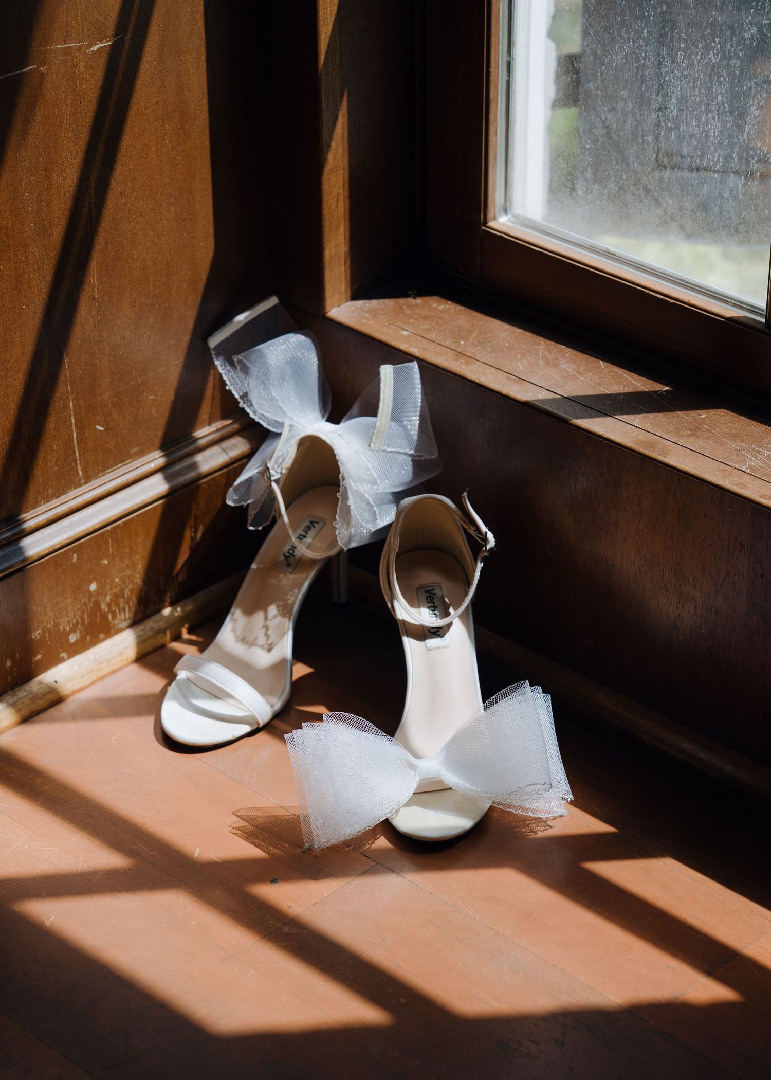 White bridal heels with tulle bow detail placed on wooden windowsill in warm sunlight.