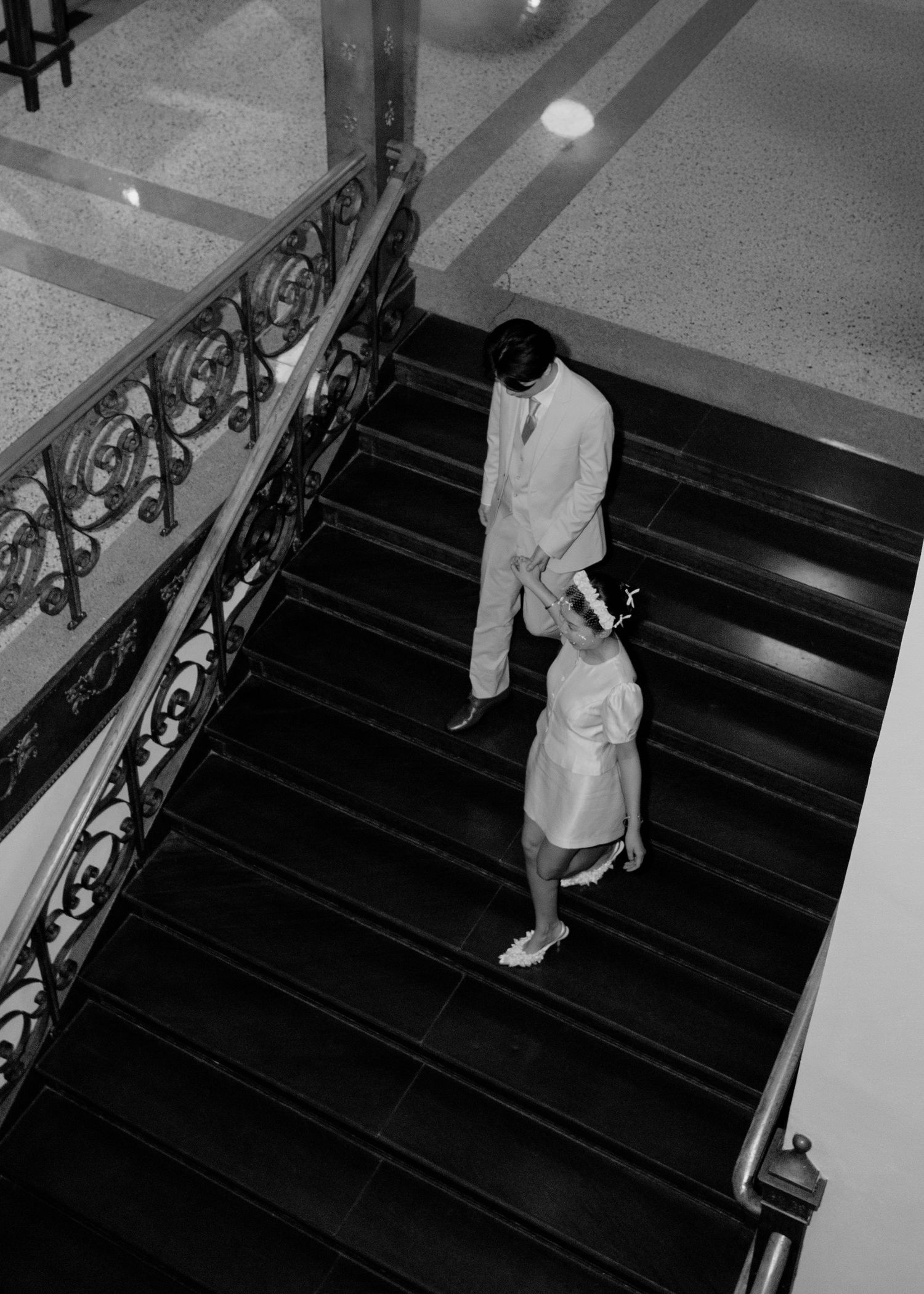 Black and white aerial view of a bride and groom descending an elegant staircase with ornate railings.