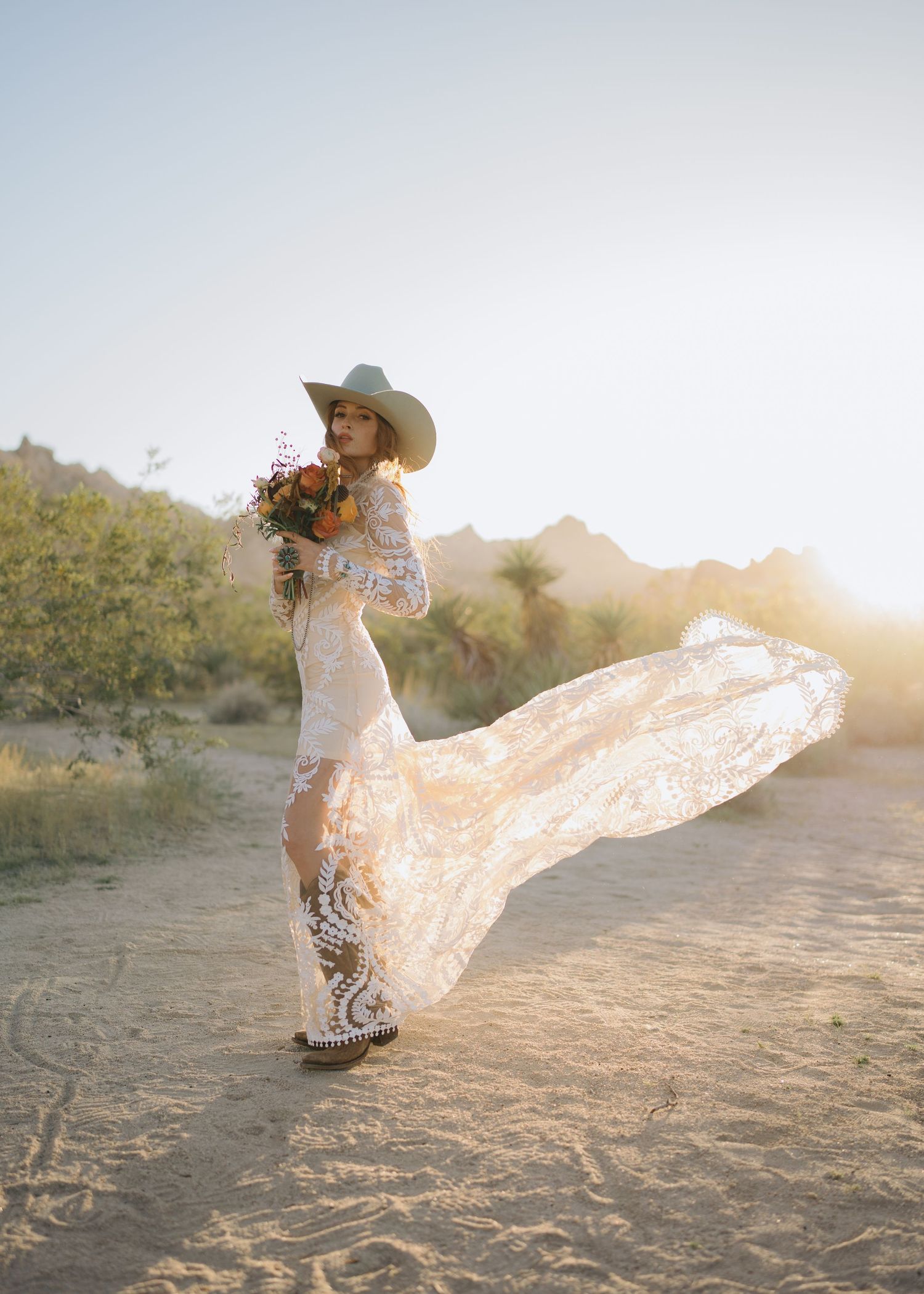 Boho bride in lace gown and cowboy hat with flowing train walking in sunlit desert landscape.