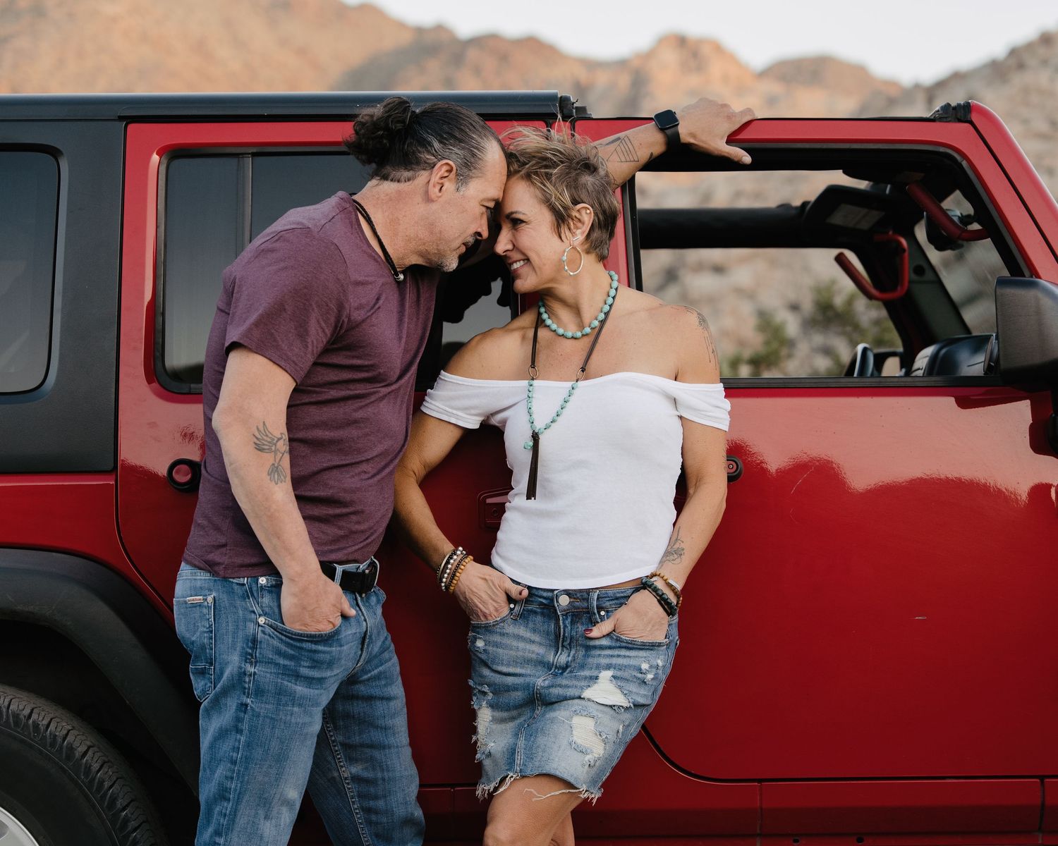 Romantic couple leaning close together against a red Jeep Wrangler in a desert landscape.