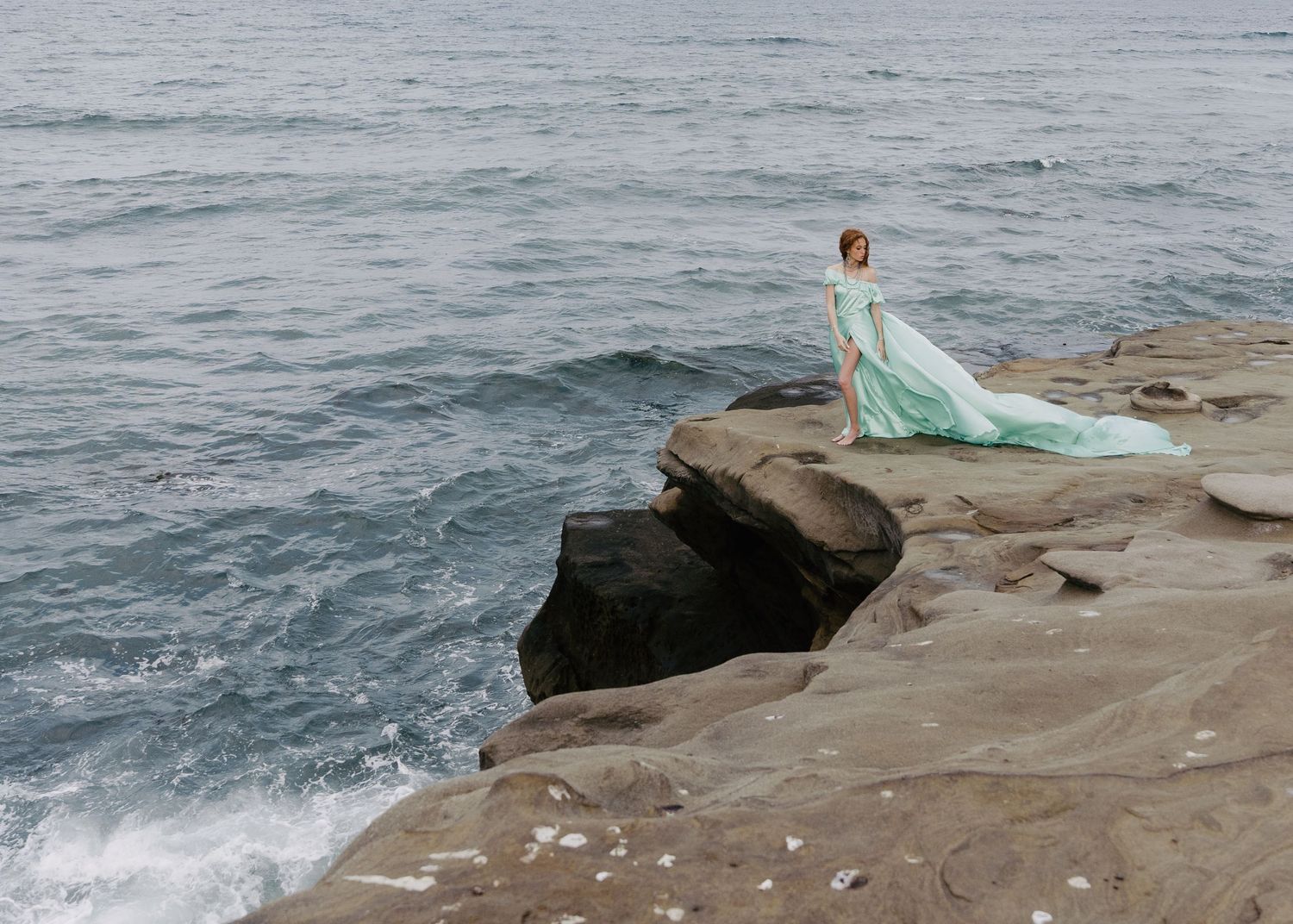 Woman in flowing mint green gown standing on dramatic rocky ocean cliffs.