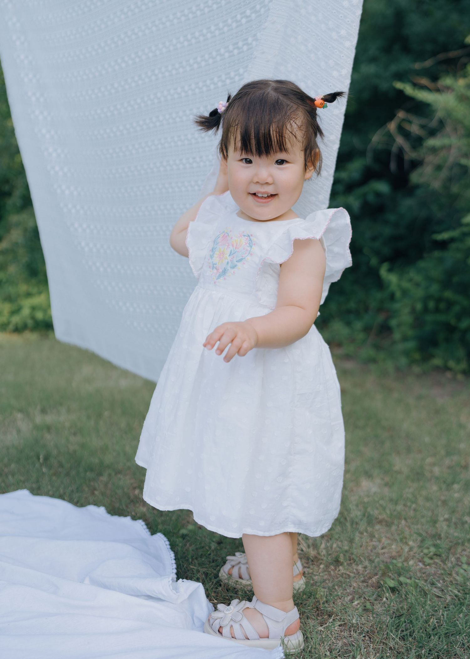 Adorable toddler girl in white ruffled dress with pigtails smiling outdoors on green lawn.