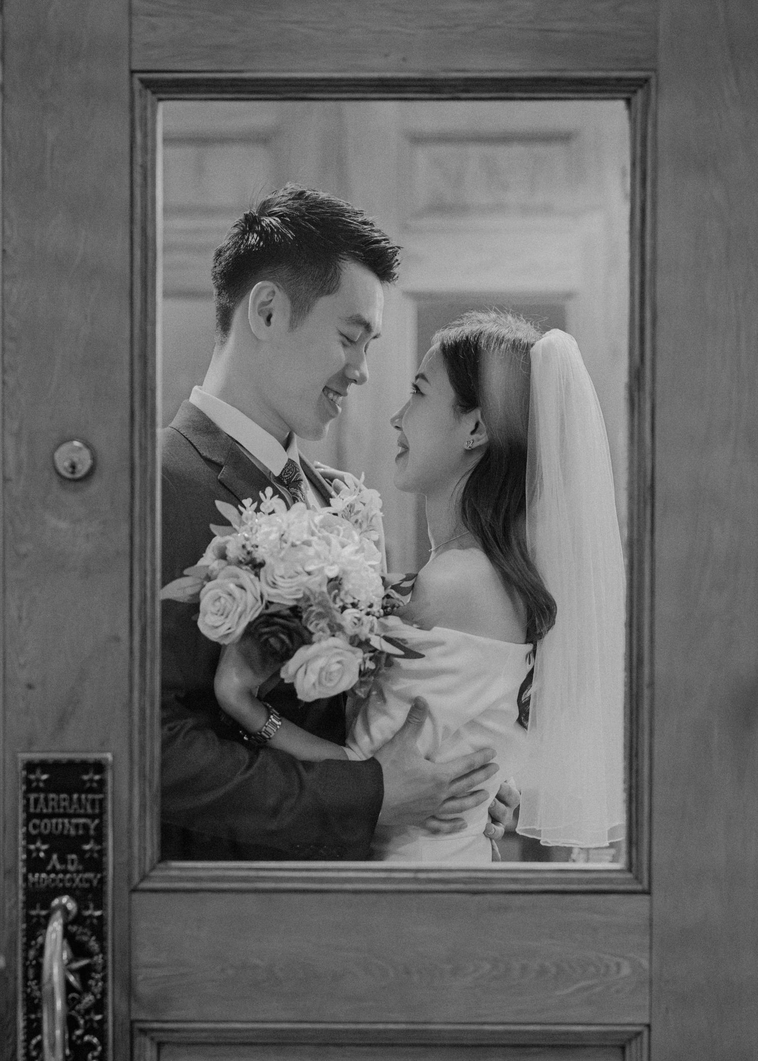 Black and white portrait of smiling Asian bride and groom embracing at Tarrant County courthouse door.
