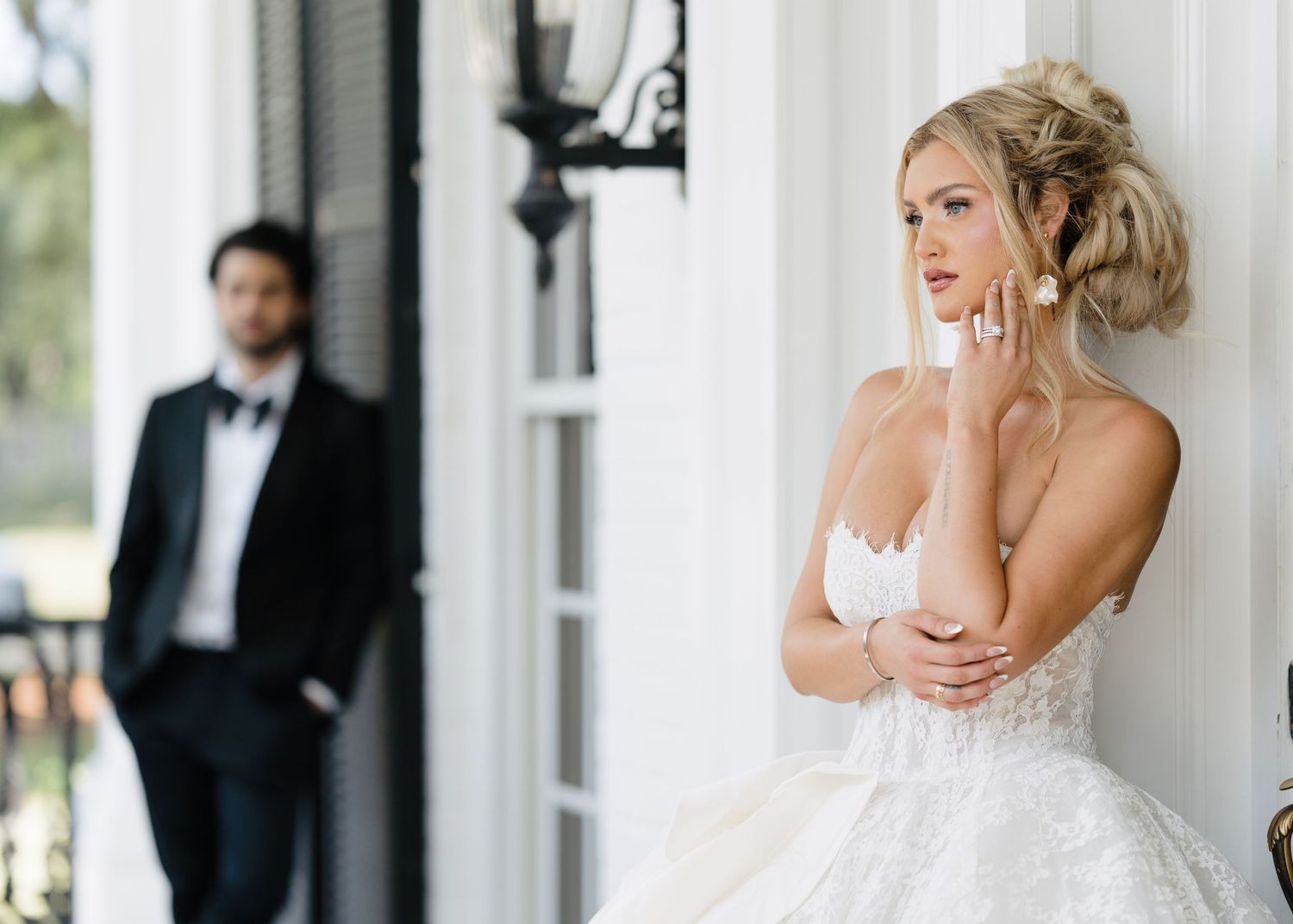Elegant bride in white lace ballgown stands on porch while groom in tuxedo stands in background.