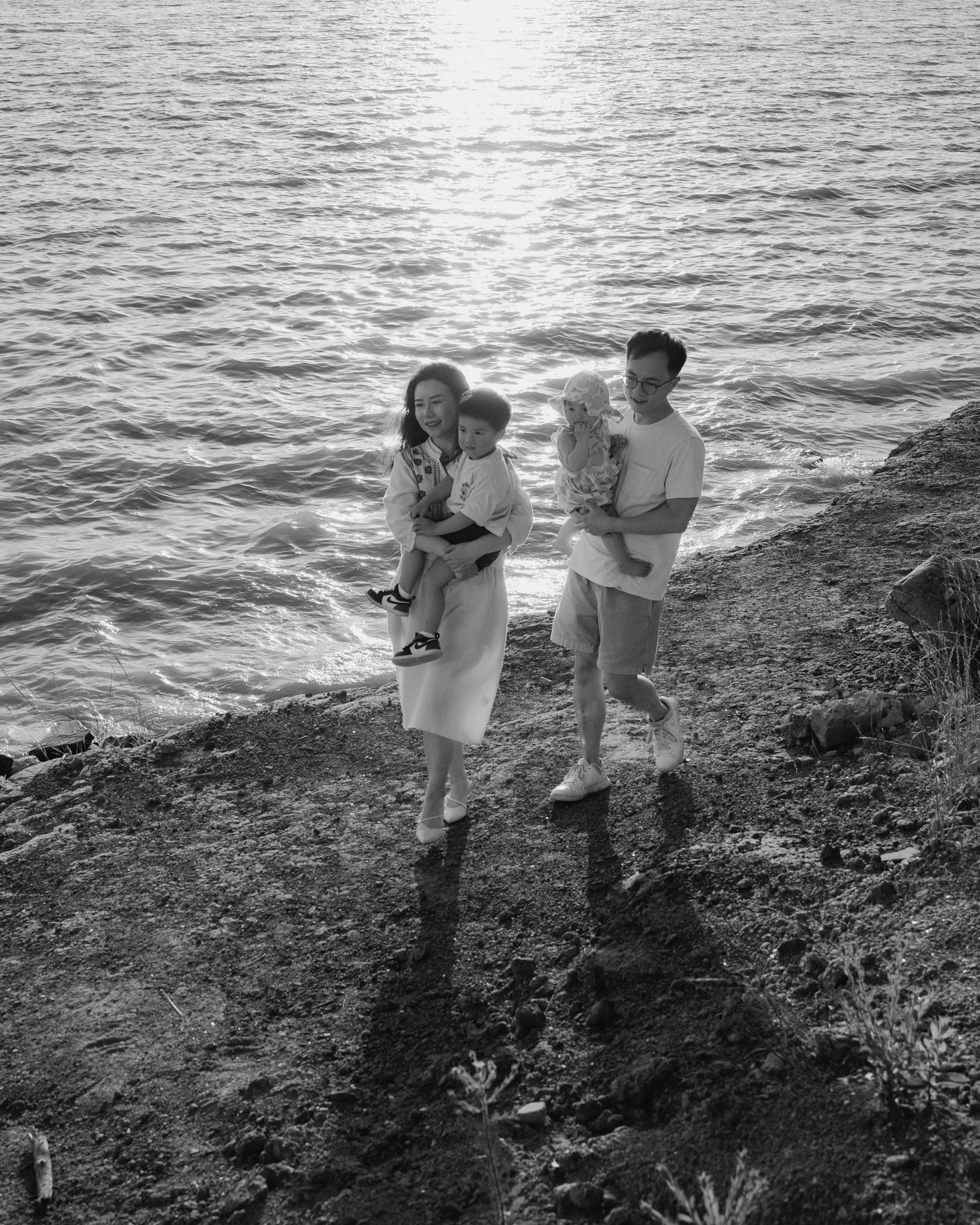 Black and white photo of a young Asian couple each holding a small child on a rocky lakeside cliff.
