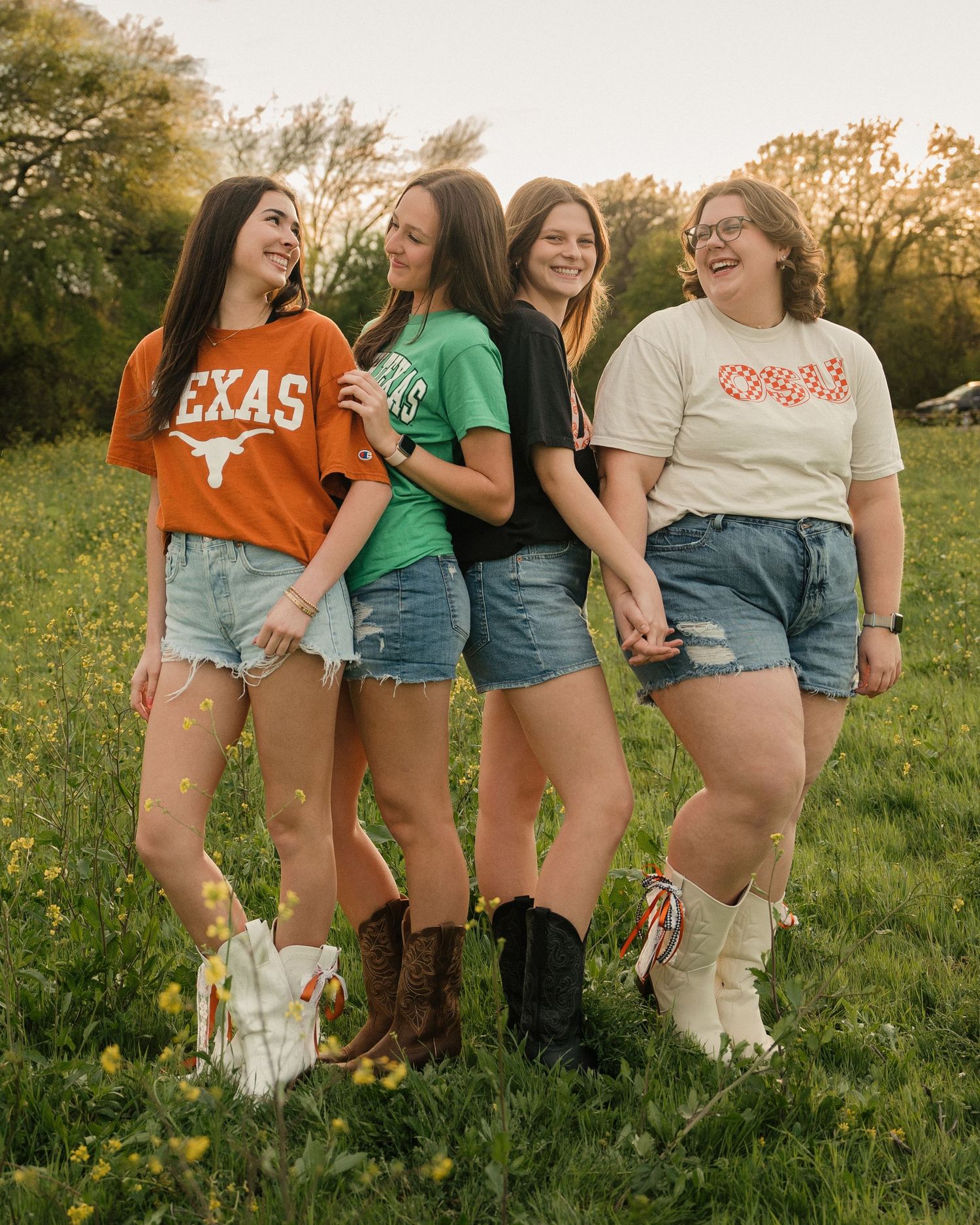 Four young women in college graphic tees and denim shorts laugh together in a green meadow.