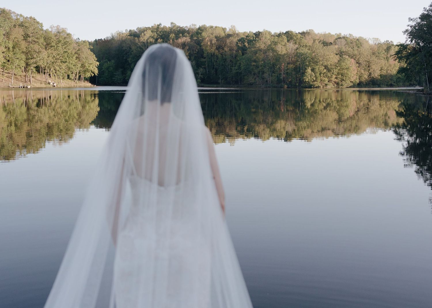 Bride in white gown and veil stands with back turned overlooking a calm reflective lake.