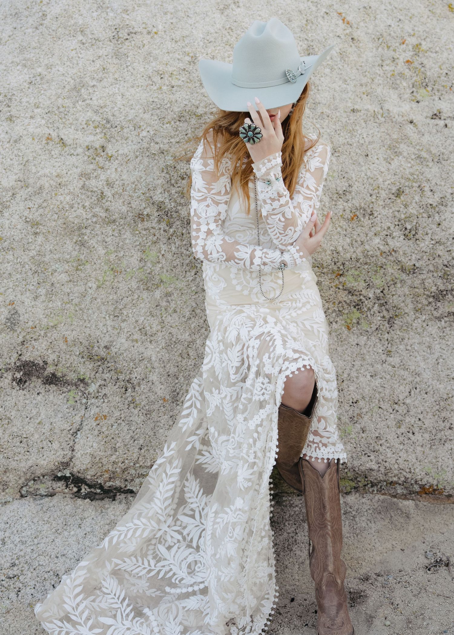 Boho bride in floral lace dress and cowboy hat wearing brown boots against stone wall.