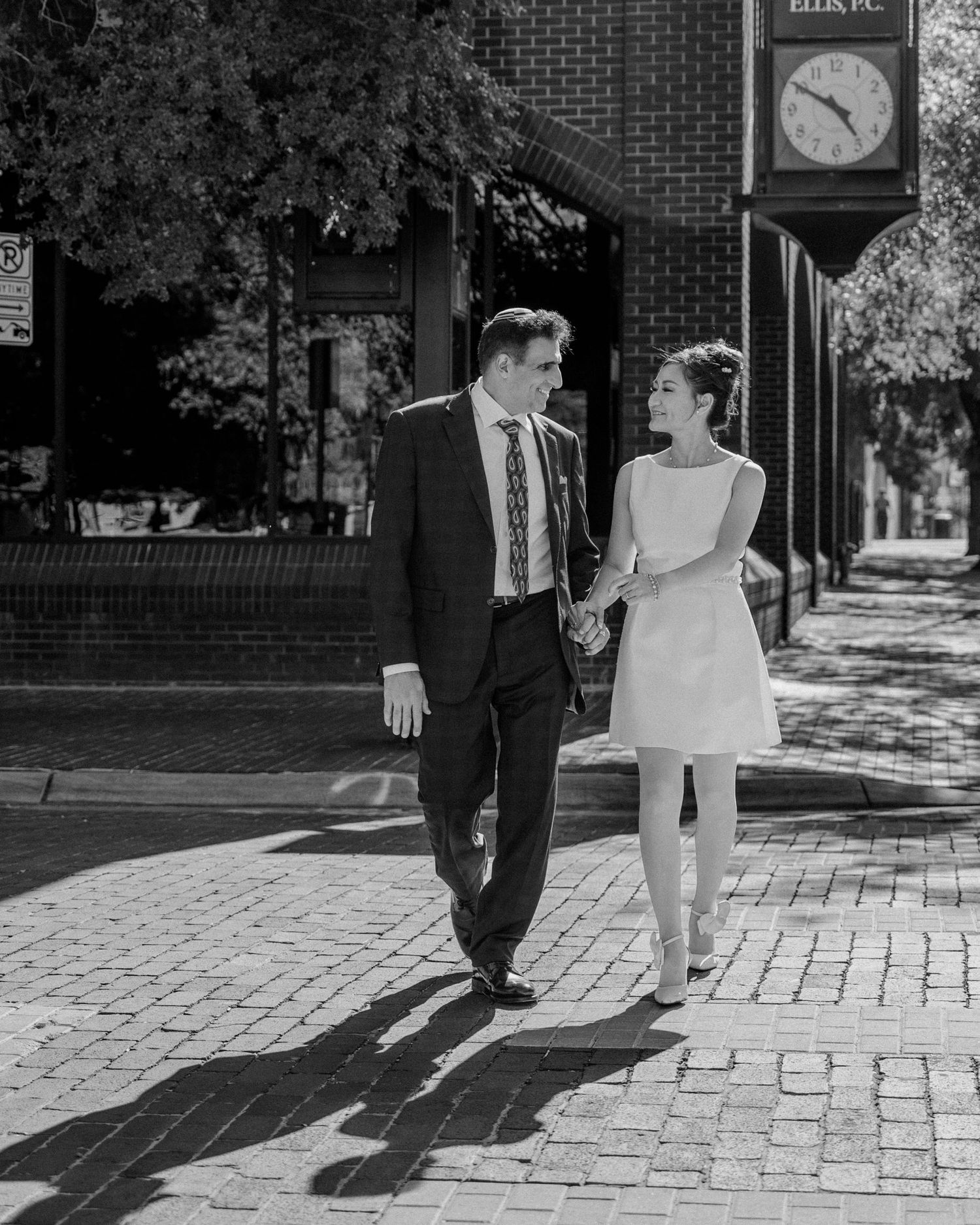 Black and white photo of couple walking hand-in-hand on sunny brick sidewalk near clock.