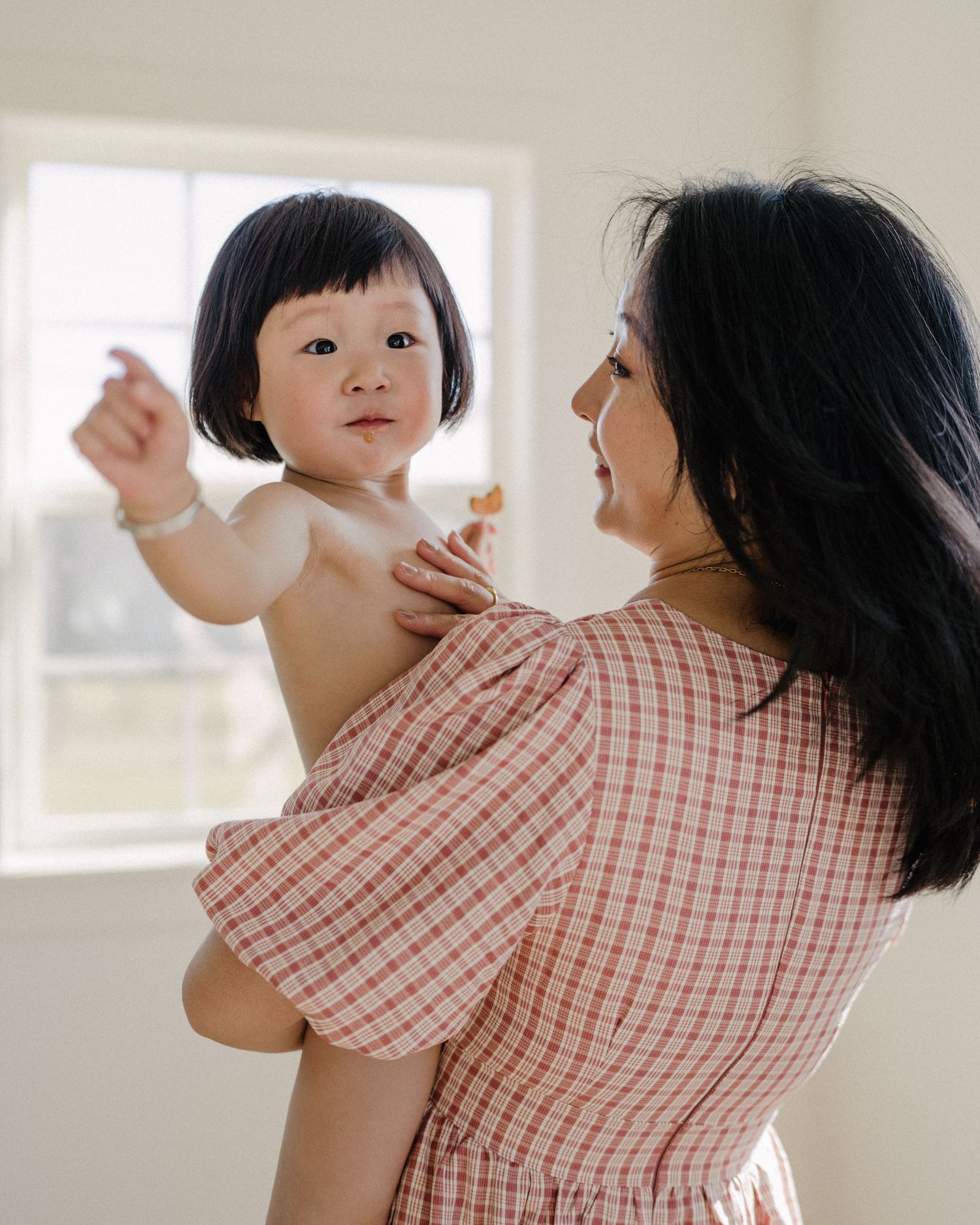 Asian mother in plaid dress holding curious baby near bright window indoors.