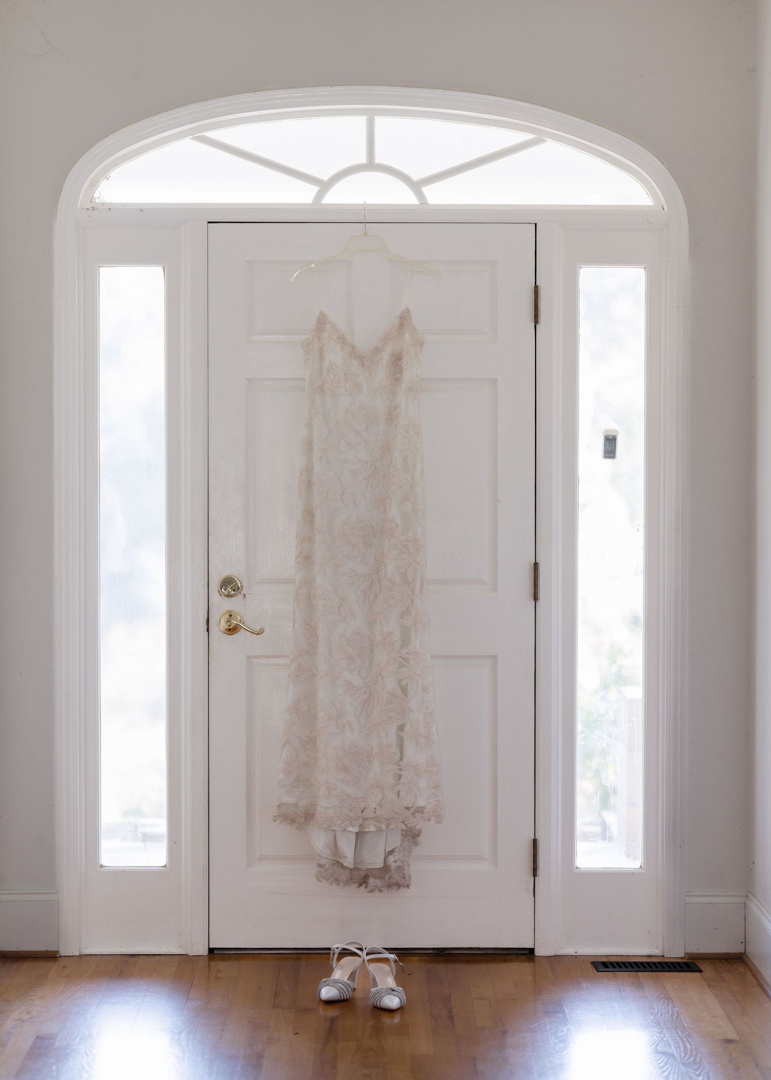 Elegant lace wedding dress hanging on white arched front door with bridal heels below.