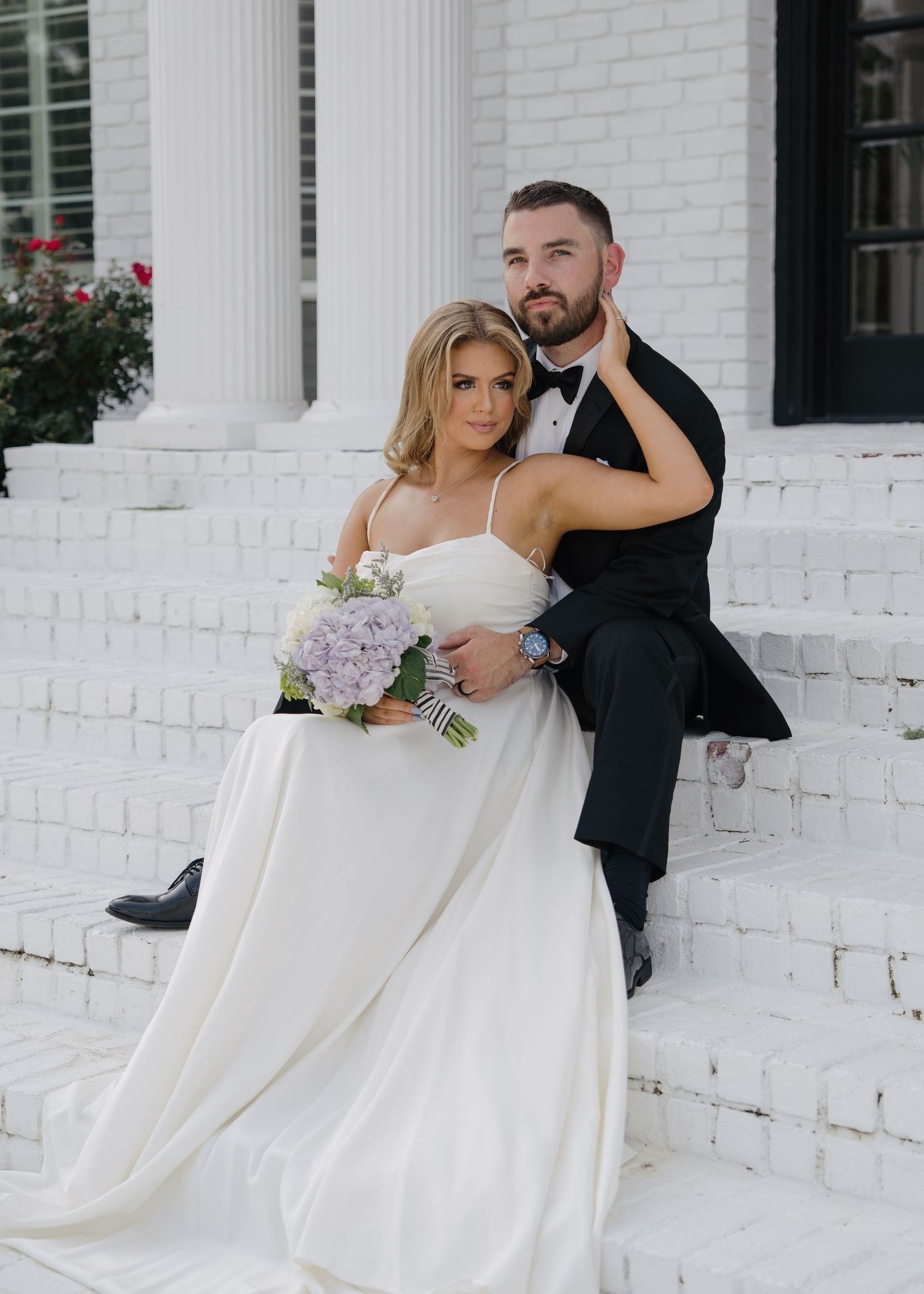 Bride in white gown with hydrangea bouquet and groom in tuxedo sitting on white steps.
