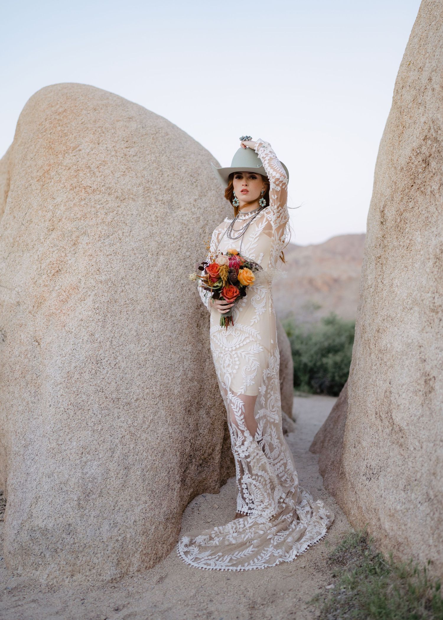 Elegant bride in lace gown and hat posing between large boulders in desert setting with bouquet.