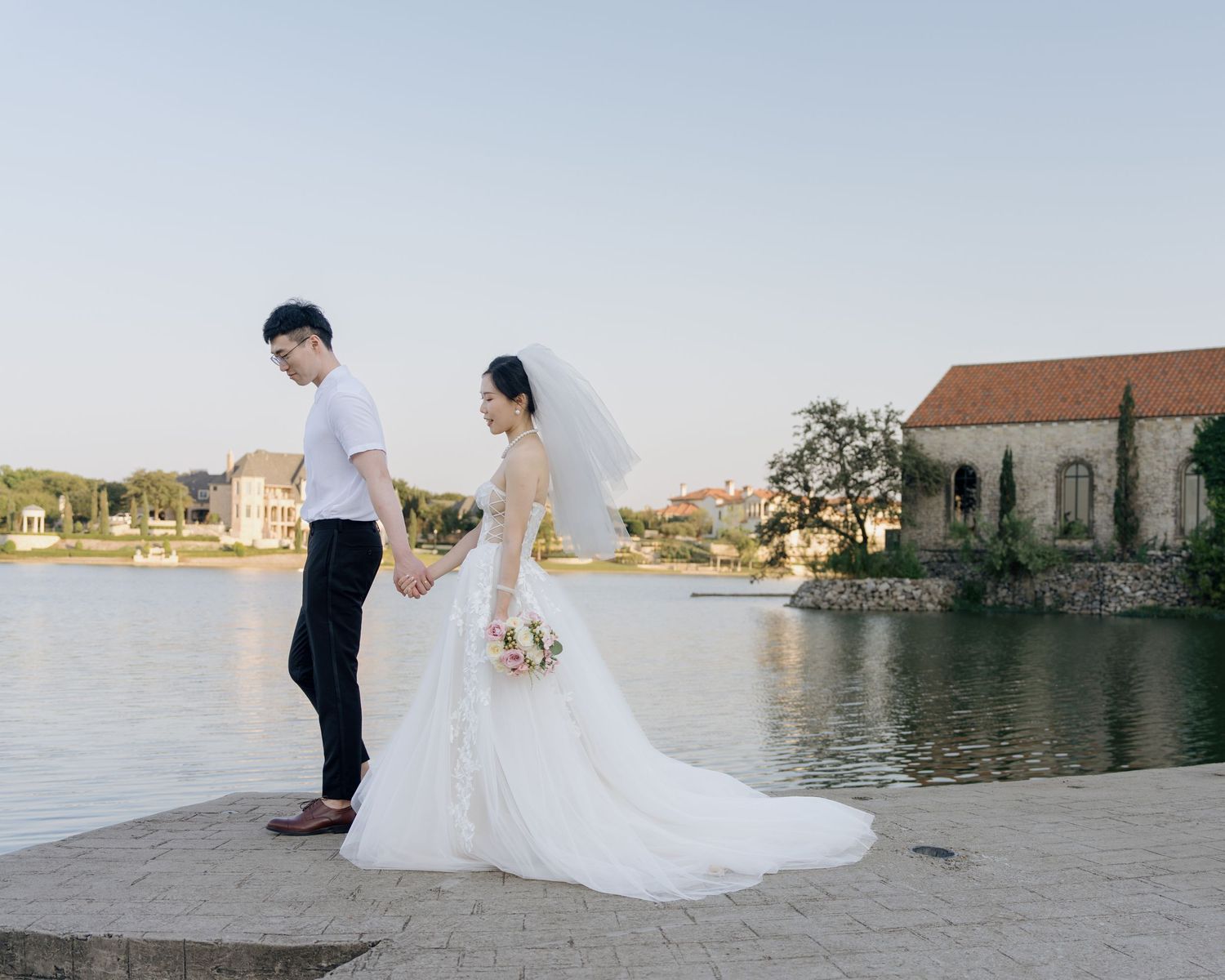 Bride and groom holding hands by a serene lakeside with a charming chapel in the background.
