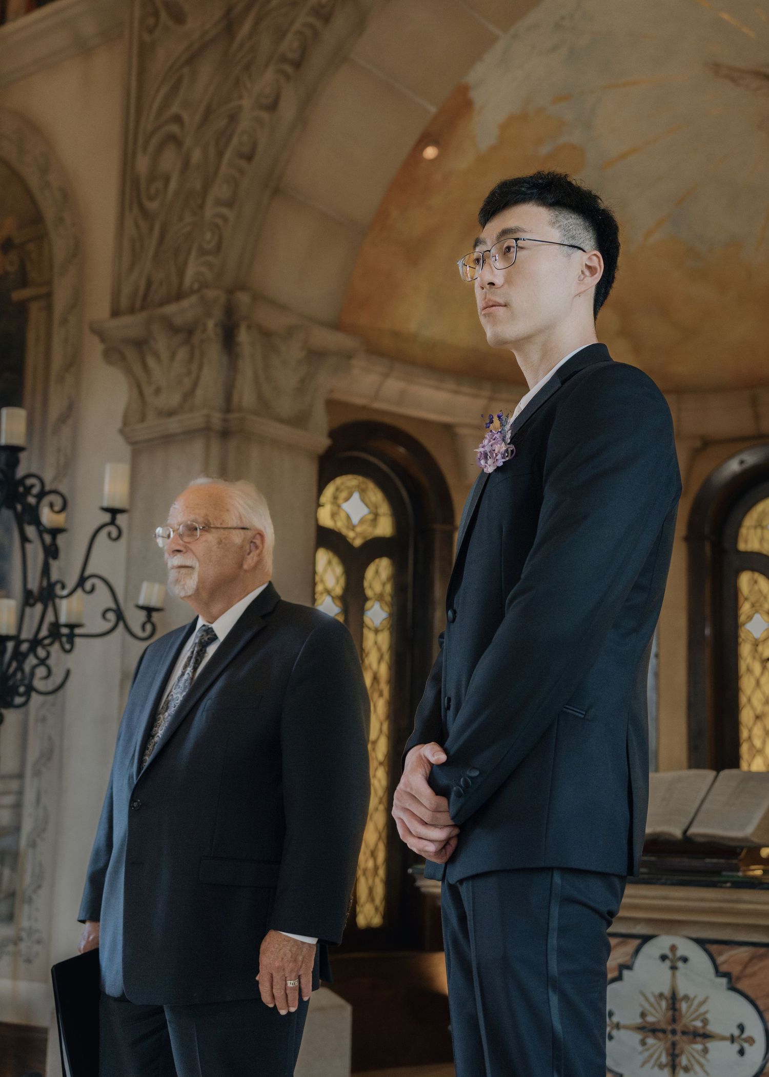 Tall groom in black suit stands with an older gentleman inside an ornate church before the ceremony.