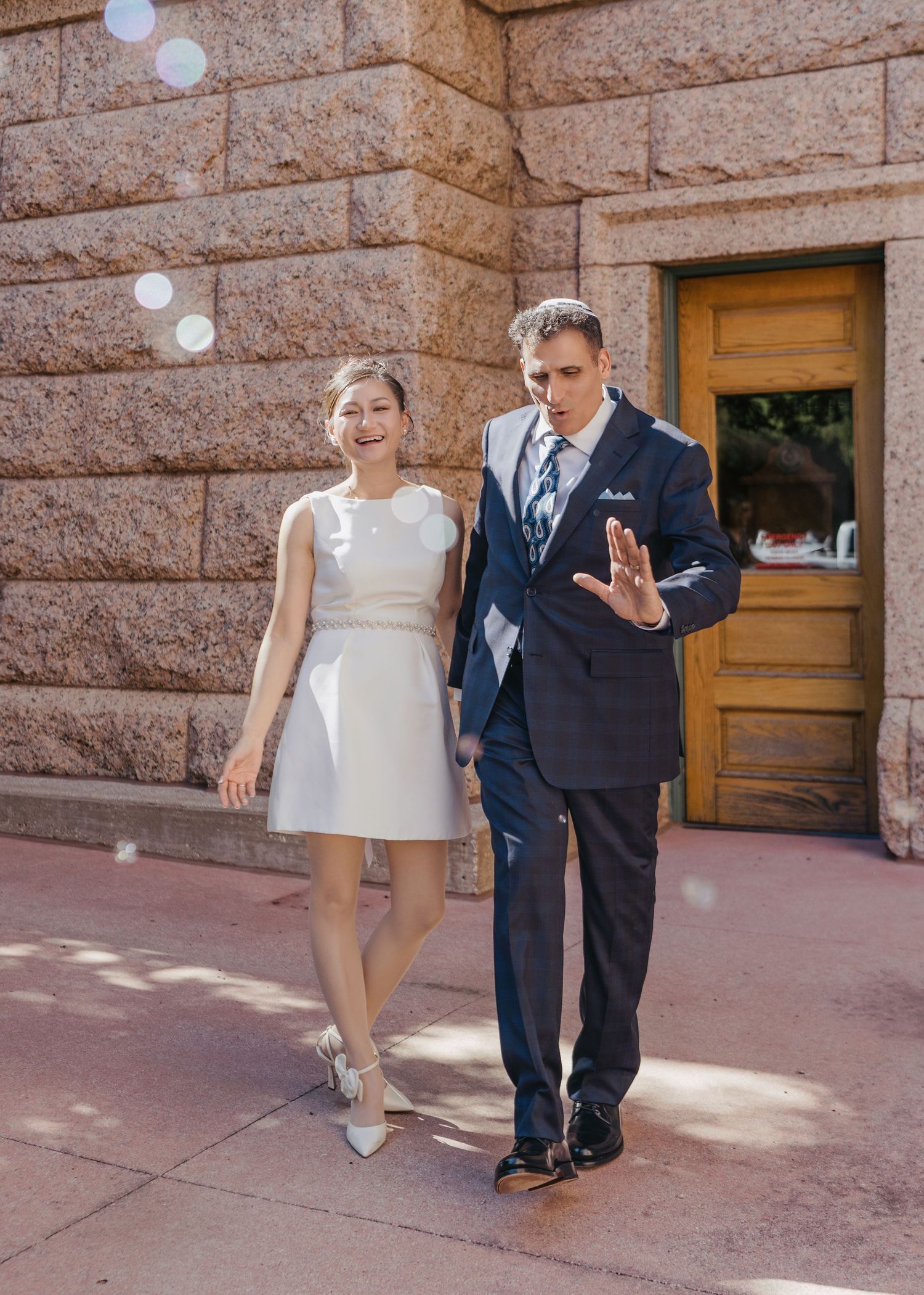 Couple laughing and walking outside historic stone building on sunny wedding day.
