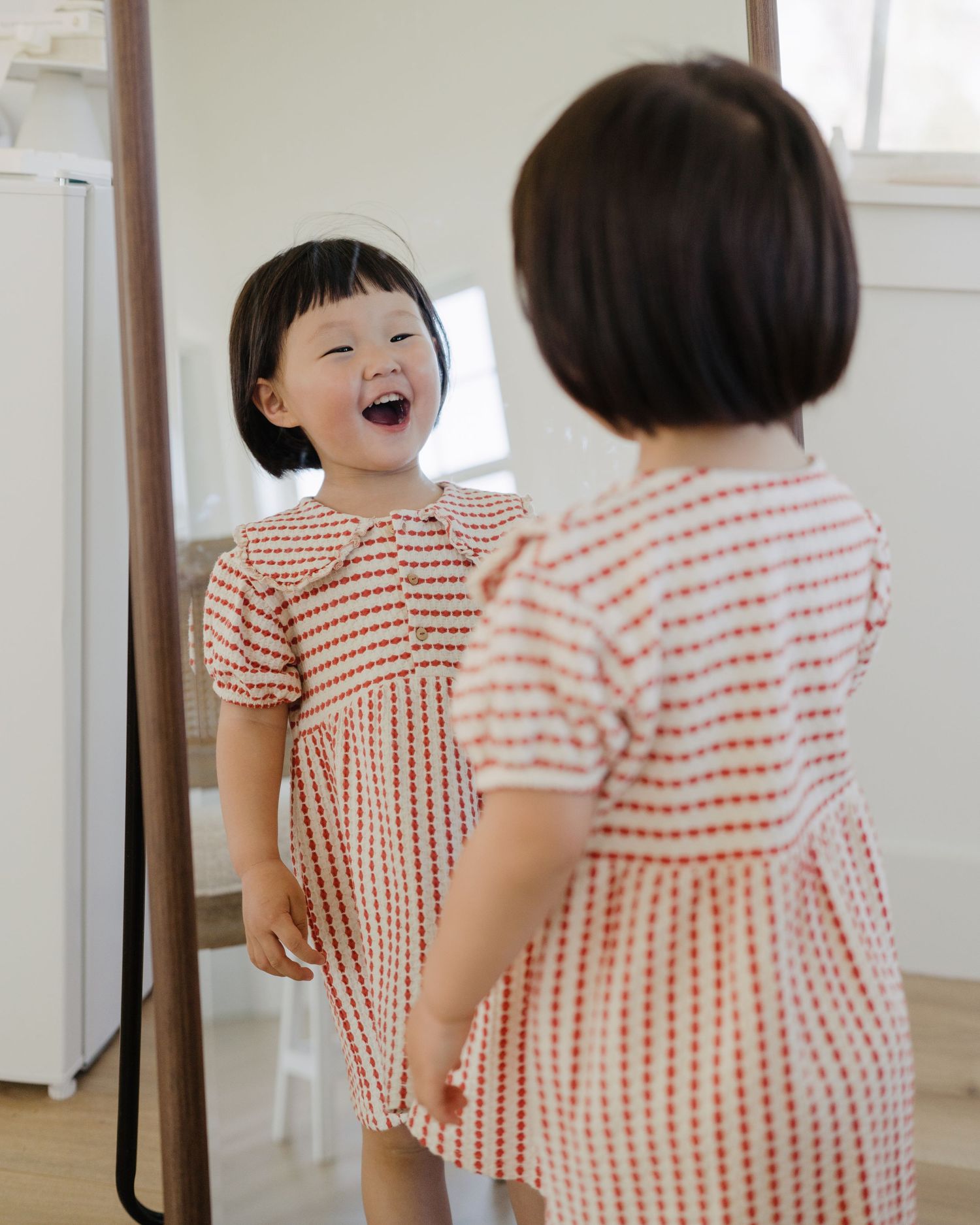 A delighted toddler girl in a red and white smocked dress laughs at her reflection in a tall mirror.
