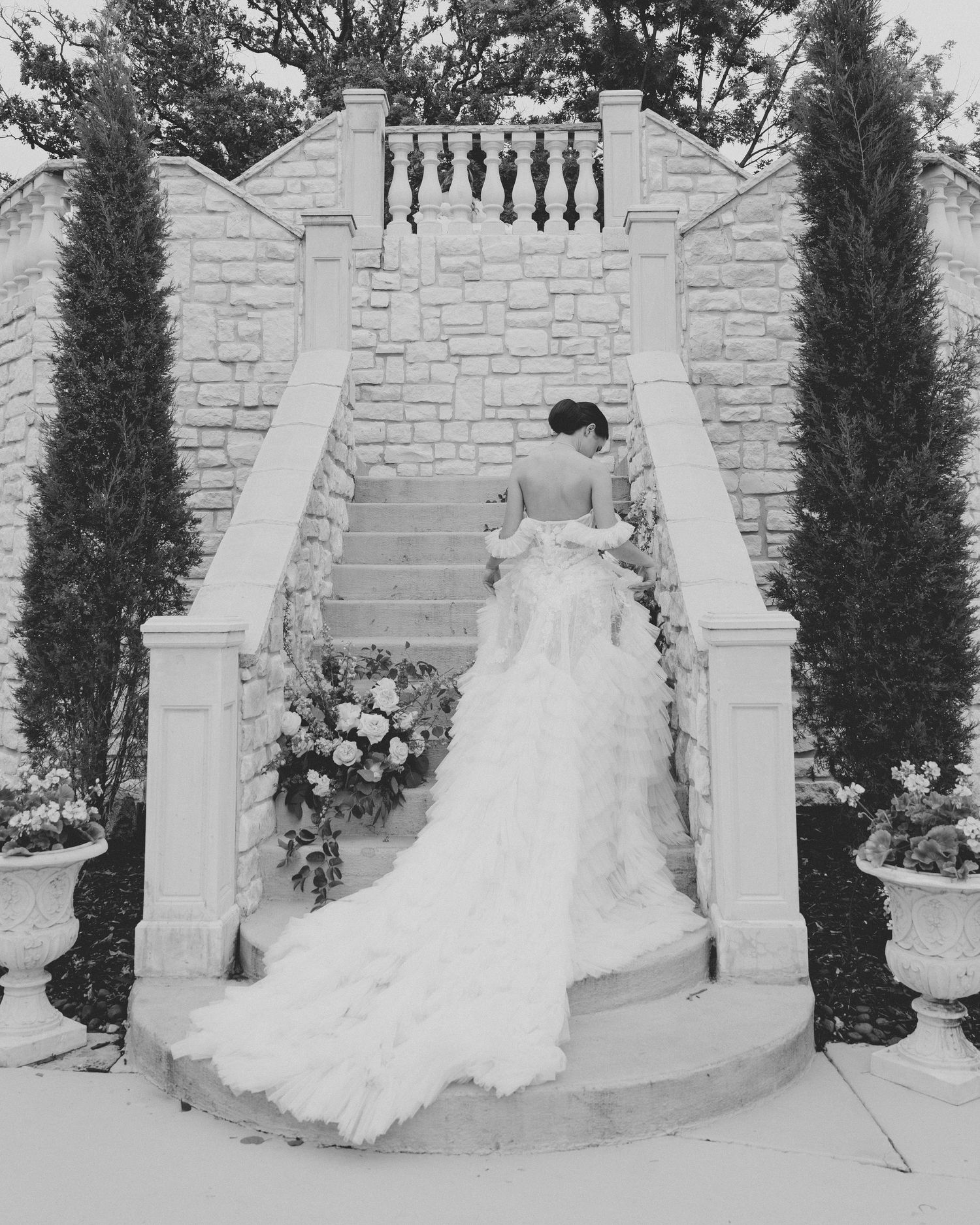 Black and white photo of bride in ruffled gown ascending stone staircase flanked by cypress trees.