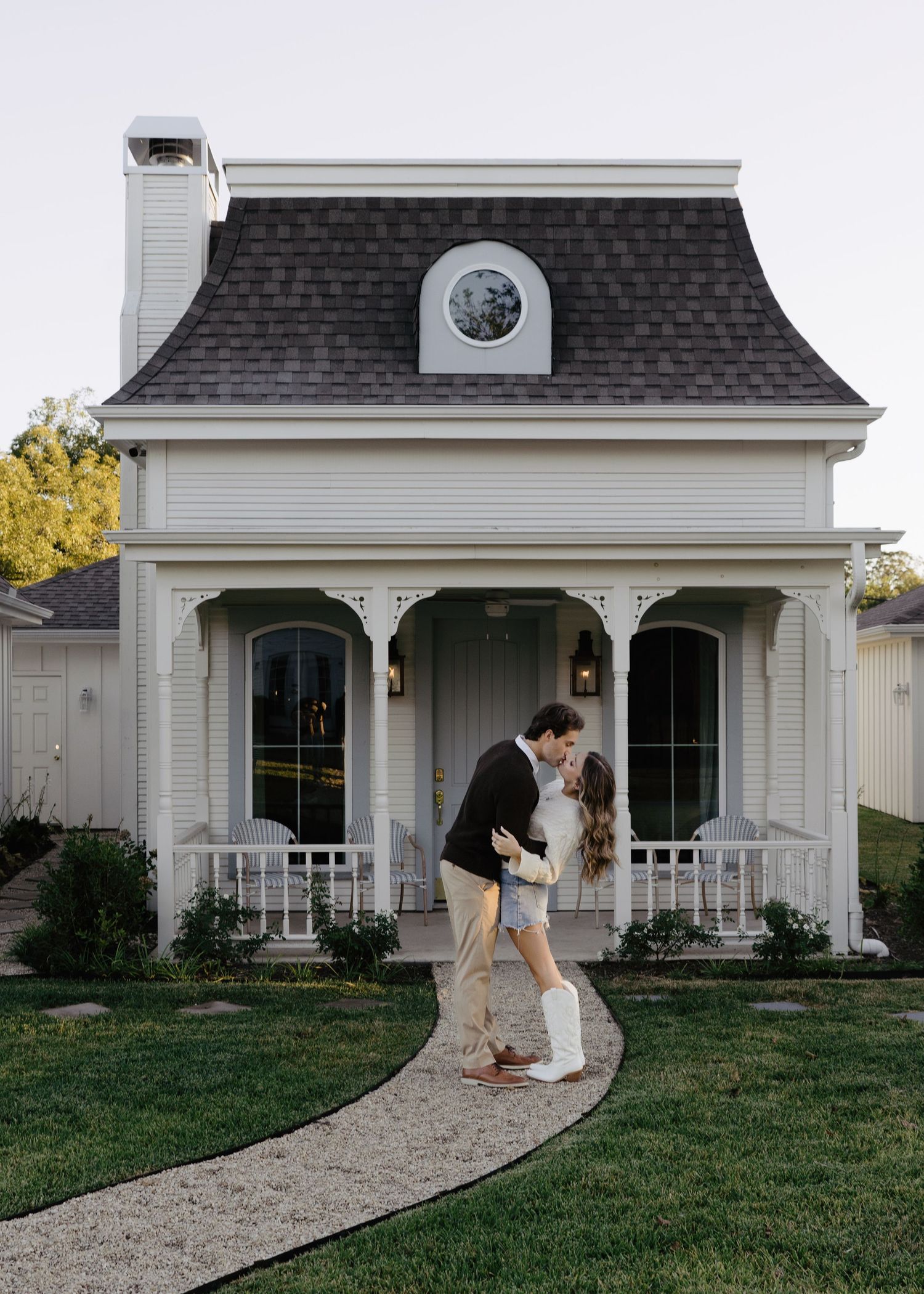 Couple sharing a kiss in front of a beautiful white Victorian-style cottage at dusk.