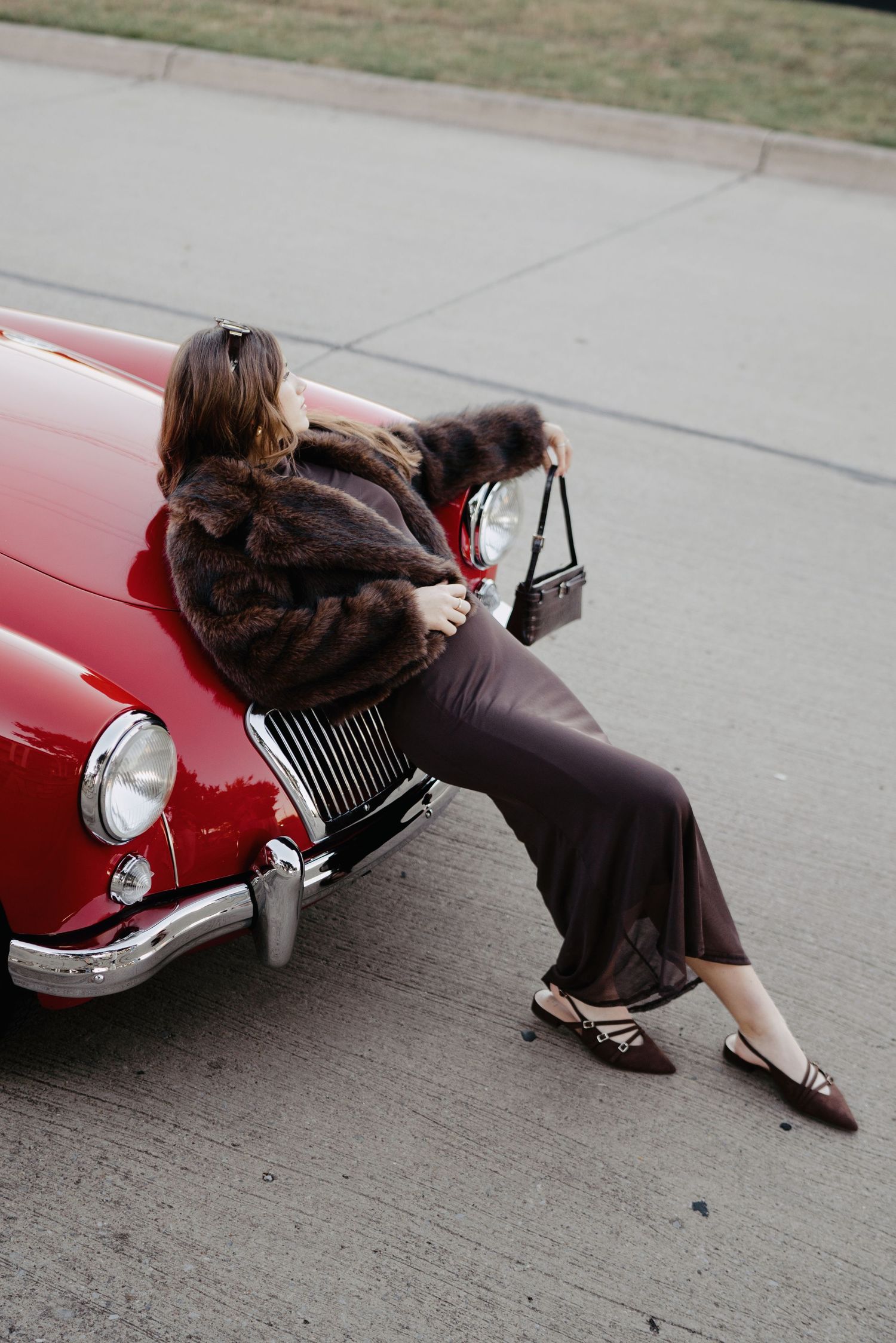 Woman in brown fur coat and maxi skirt sitting on the hood of a classic red vintage car.