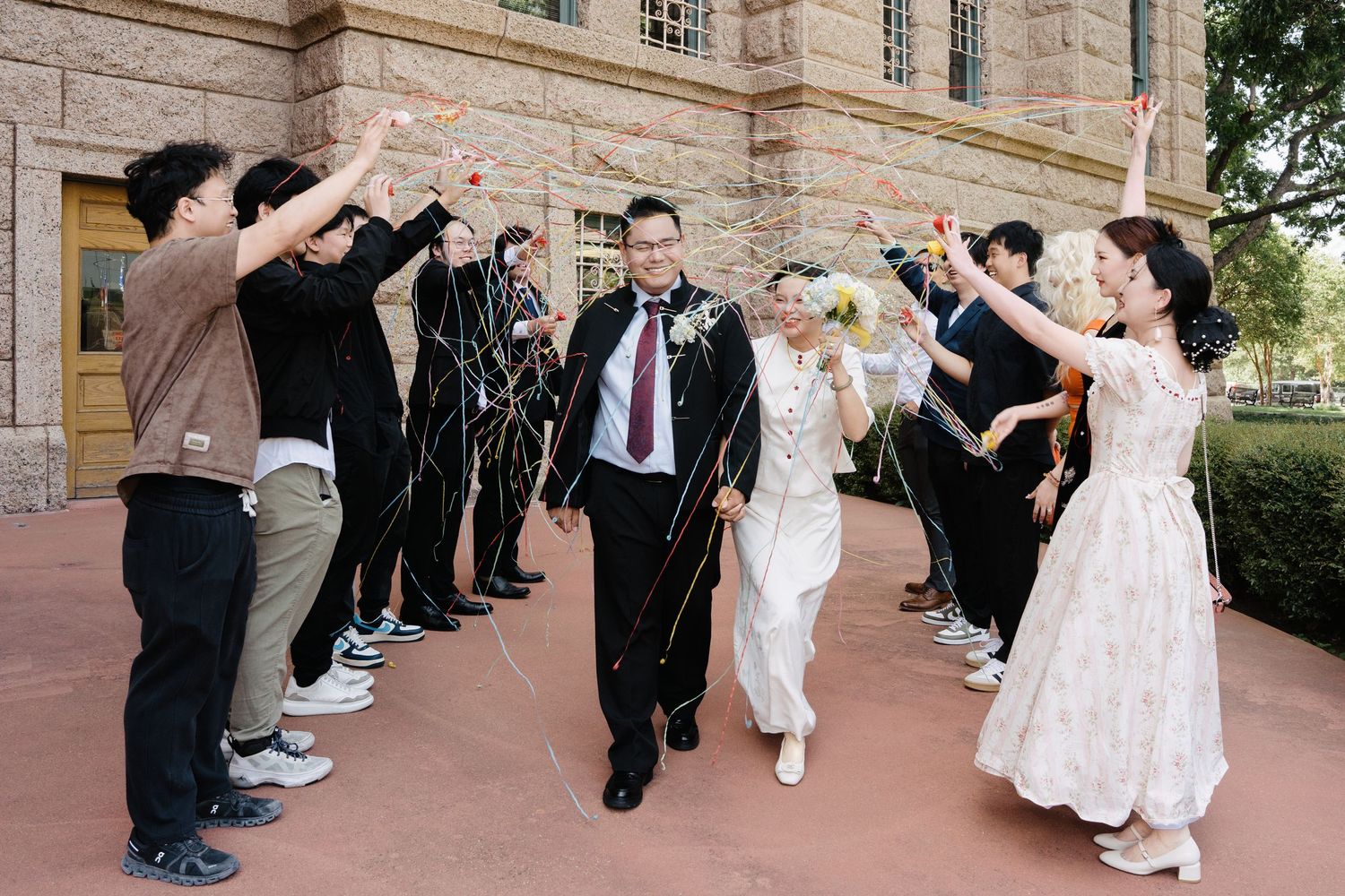 Newlywed couple walks through a celebratory arch of streamers with wedding party outside a brick building.